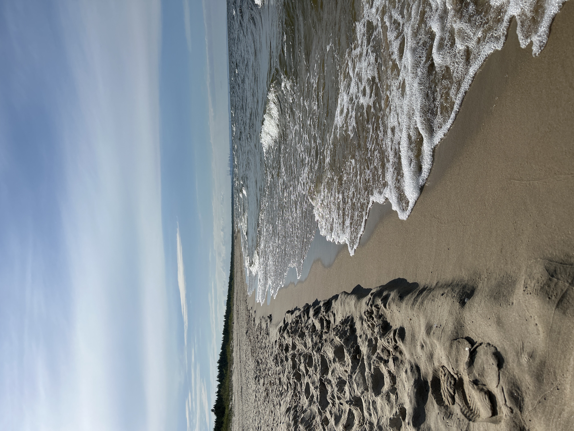 The image depicts a beach scene in Gdańsk, Poland. Waves are washing onto a sandy shore. In the foreground, there are footprints in the sand. A line of trees is visible along the coastline. The sky is blue with scattered clouds.