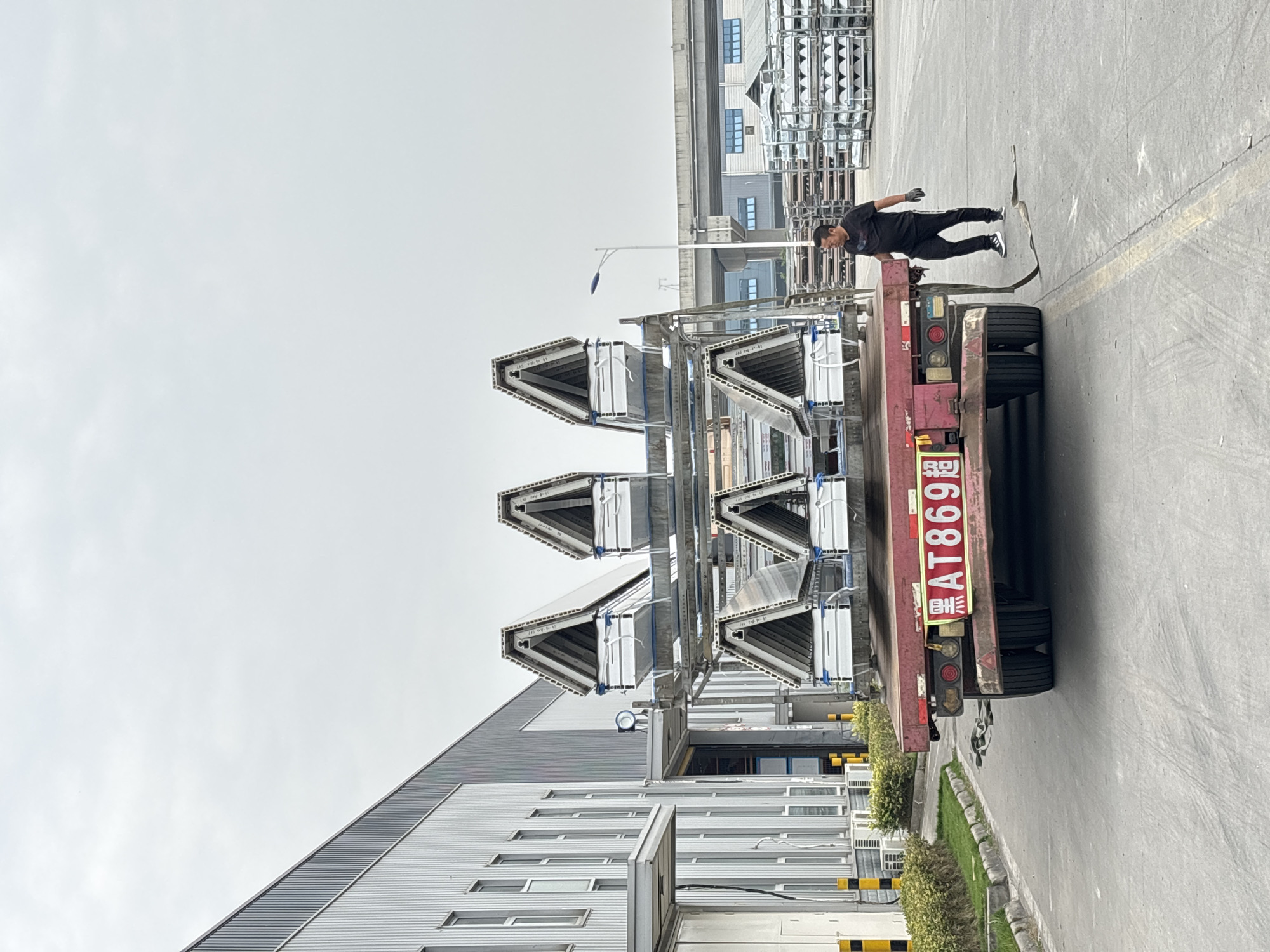 A wide-angle shot shows a person standing on a flatbed truck loaded with triangular metal structures, likely in Chengdu, China. The truck is parked on an asphalt surface with buildings in the background and alongside a narrow strip of green landscaping.
The truck has a red license plate that reads "渝AT869超". The metal structures on the flatbed appear to be prefabricated building sections. The person standing on the truck is dressed in dark clothing. The sky is overcast.