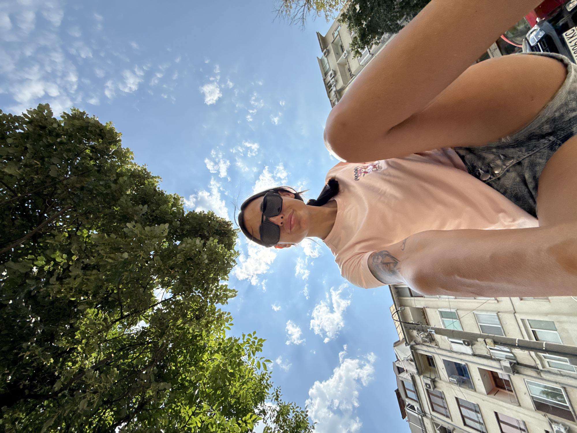 The image depicts a young woman wearing a pink t-shirt and denim shorts, posing for a selfie outdoors. She has dark hair pulled back and is wearing large black sunglasses. Her left arm, visible on the left side of the frame, has a tattoo. The background shows a bright blue sky with scattered white clouds, lush green tree foliage overhead, and a light-colored apartment building. The perspective suggests the photo was taken from a low angle, looking upwards. The license plate of a parked car, "BG 0061 KC", is partially visible at the bottom right. The architectural style of the building and the license plate format are indicative of a location in Bulgaria, specifically the city of Pleven.