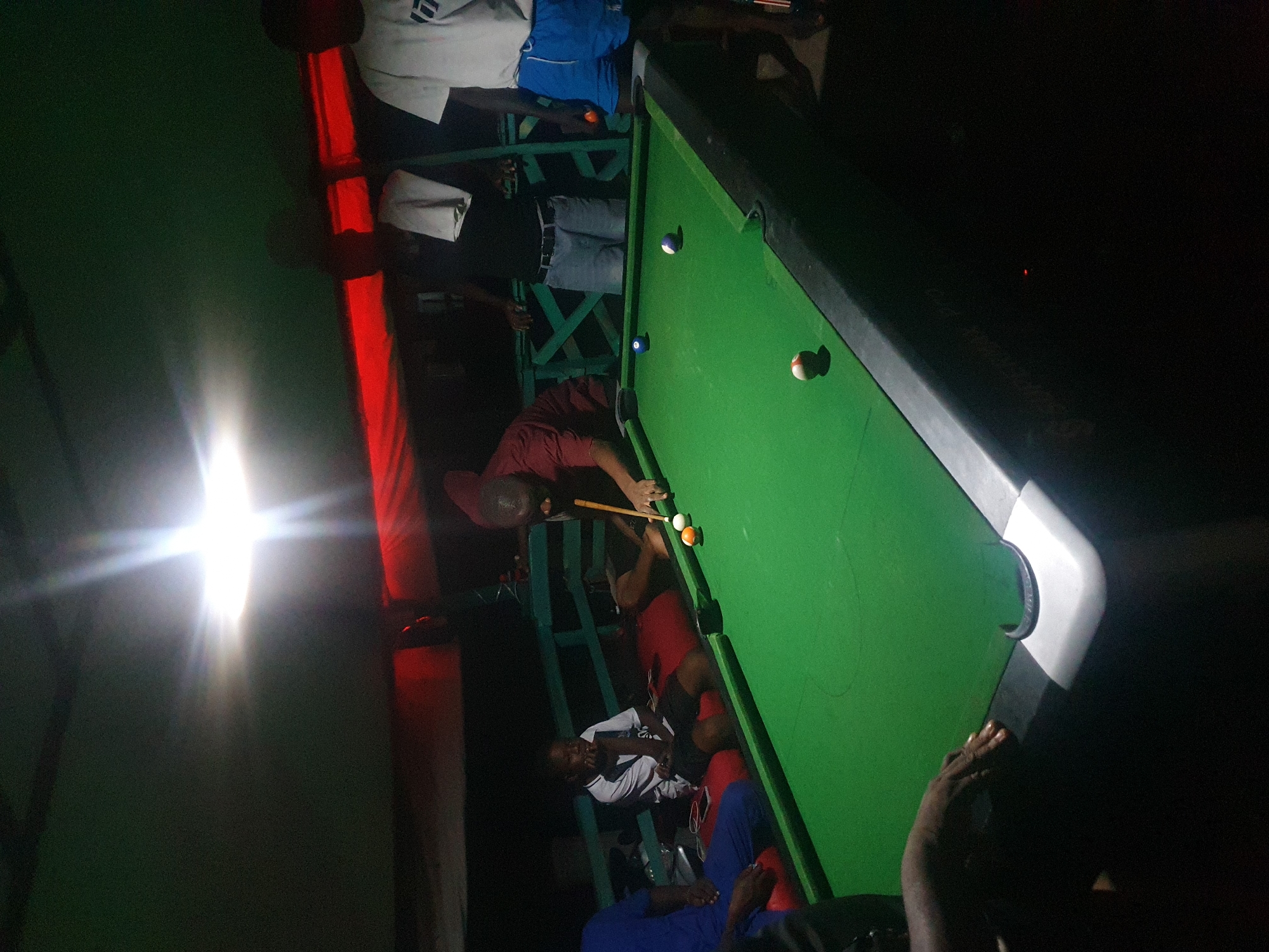 The image depicts an outdoor setting, possibly a bar or recreational area, at night. A pool table with green felt is the central focus, with several people gathered around it. In the foreground, a person is in the process of playing pool, holding a cue stick and aiming at a ball. There are multiple billiard balls on the table, including a red and white striped ball, a solid orange ball, and a white cue ball.
Several individuals are visible in the background and around the table. One person is seated on a red cushion, appearing to be watching the game. Another individual in a white t-shirt and blue shorts stands to the right, holding an orange billiard ball. Other people are partially visible, suggesting a social gathering. The environment appears to be covered by a green tarp or roof, and a bright light source, possibly a spotlight, illuminates the scene from above, creating strong shadows and highlights. The setting appears to be a dimly lit, open-air space. The presence of a "SUPERMAX PRO" inscription on the pool table suggests the brand of the table. The image context implies the location is Lokoja, Nigeria, indicating a local leisure spot.
