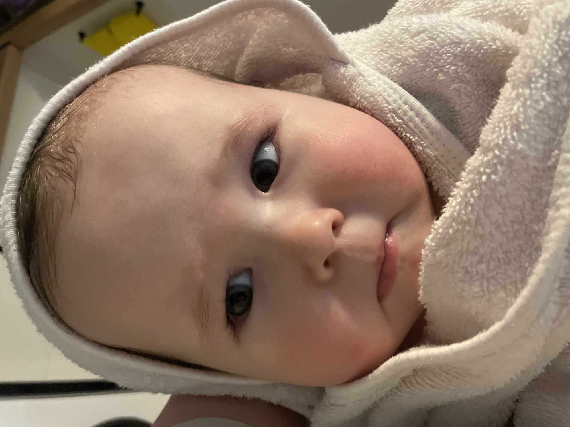 A close-up shot depicts a baby wrapped in a white towel. The baby's face is centered, with a slight tilt. The skin is fair, with a healthy pink blush on the cheeks. The eyes are dark. The towel covers the baby's head, creating a hood-like effect. There is no indication this photo was taken in Chaikovsky, Russia.