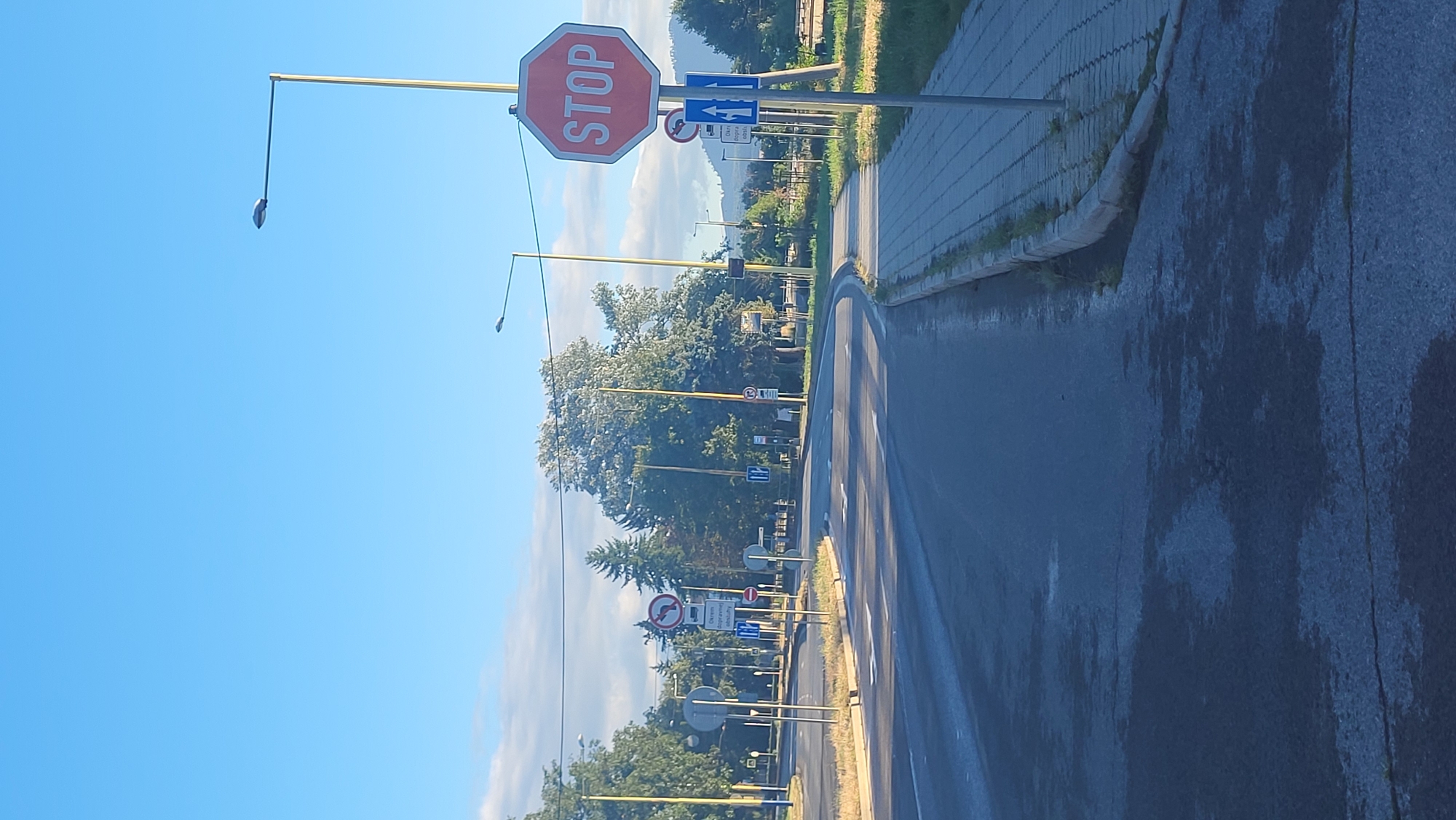 The image shows a street scene in Prešov, Slovakia, on a clear day with a blue sky and some clouds. A road curves to the right, with a sidewalk on the right side of the road. There are several traffic signs visible, including a prominent red octagonal "STOP" sign. Other signs include a circular sign with a red border and a diagonal red bar, a circular sign with a red border and a vehicle symbol, and blue rectangular signs with white arrows indicating traffic direction. Streetlights are present along the road. Trees and some buildings are visible in the background. The road surface appears wet in some areas.