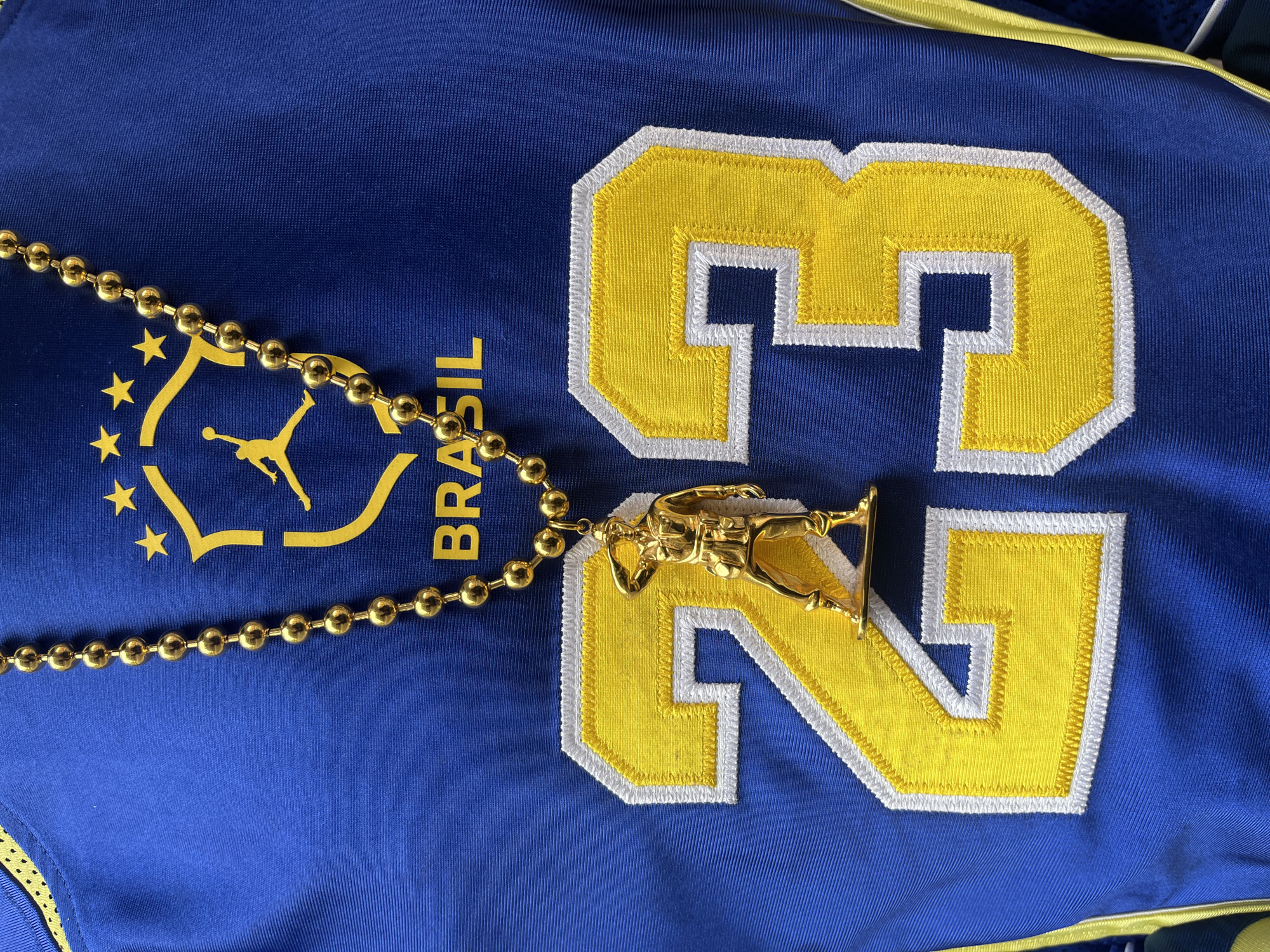 A close-up shot features a blue fabric surface with yellow and white appliqué detailing, likely a sports jersey. The appliqué forms the number "23" and the word "BRASIL", along with a logo depicting a stylized figure in silhouette surrounded by stars. A gold-colored ball chain necklace is draped across the fabric. A gold-colored statuette, seemingly of a military figure, lies atop the appliqué number "23." The lighting is bright and appears to be artificial. The location, London, United Kingdom, cannot be determined from the visual elements.