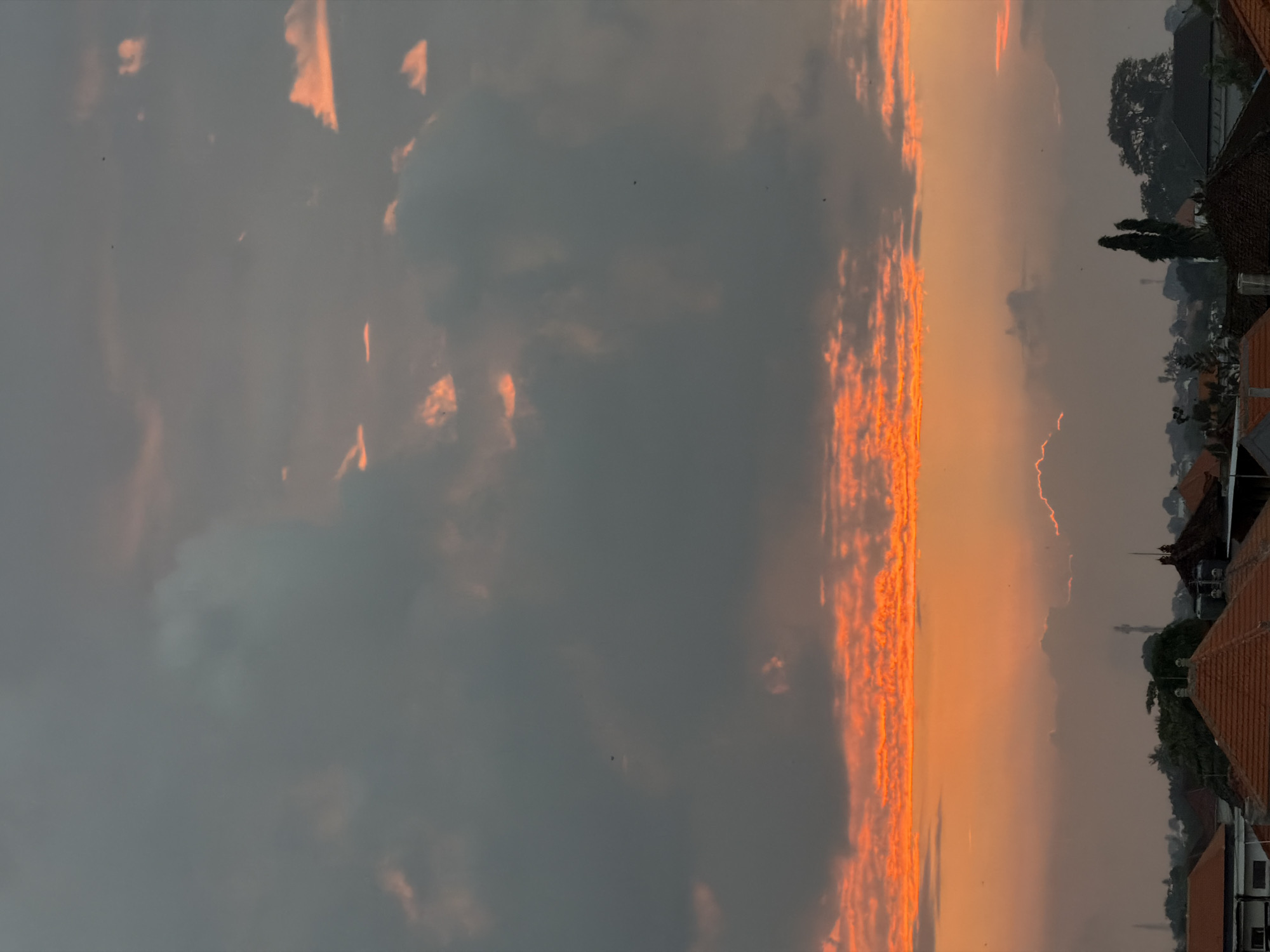 The image depicts a dusky sky over the rooftops of North Kuta, Indonesia. The sky is a dramatic canvas of dark gray clouds interspersed with vibrant streaks of orange and red, suggesting a sunset or sunrise. Scattered, small dark specks, possibly birds, are visible against the sky. Below the horizon, the silhouettes of buildings with terracotta roofs are apparent, indicative of a dense urban or suburban landscape. Trees and other foliage are also visible, adding to the texture of the scene. The overall impression is of a serene yet striking natural phenomenon occurring above a settled area.