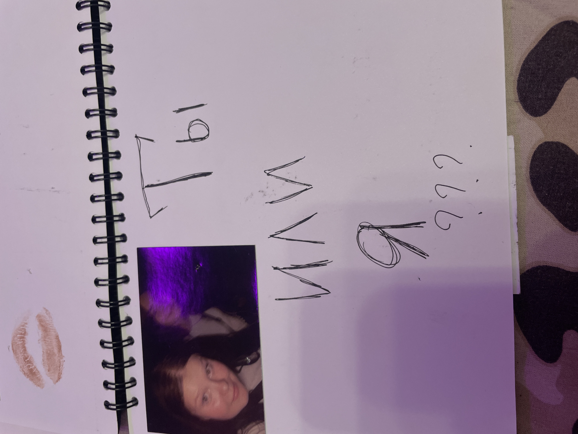 A photograph shows an open spiral-bound notebook on a surface that appears to be camouflage fabric. On the left page of the notebook, there is a lipstick imprint. To the right of the lipstick imprint, there are three lines of handwritten Russian text. The first line reads "Ты" (Ty, meaning "You"). The second line reads "или" (ili, meaning "or"). The third line reads "я" (ya, meaning "I"), followed by three question marks. There is also a small, blurry photograph inserted into the notebook, showing a young woman with dark hair. The notebook is positioned on a piece of camouflage patterned fabric. No location in Vaganovo, Russia is identifiable in this image.
