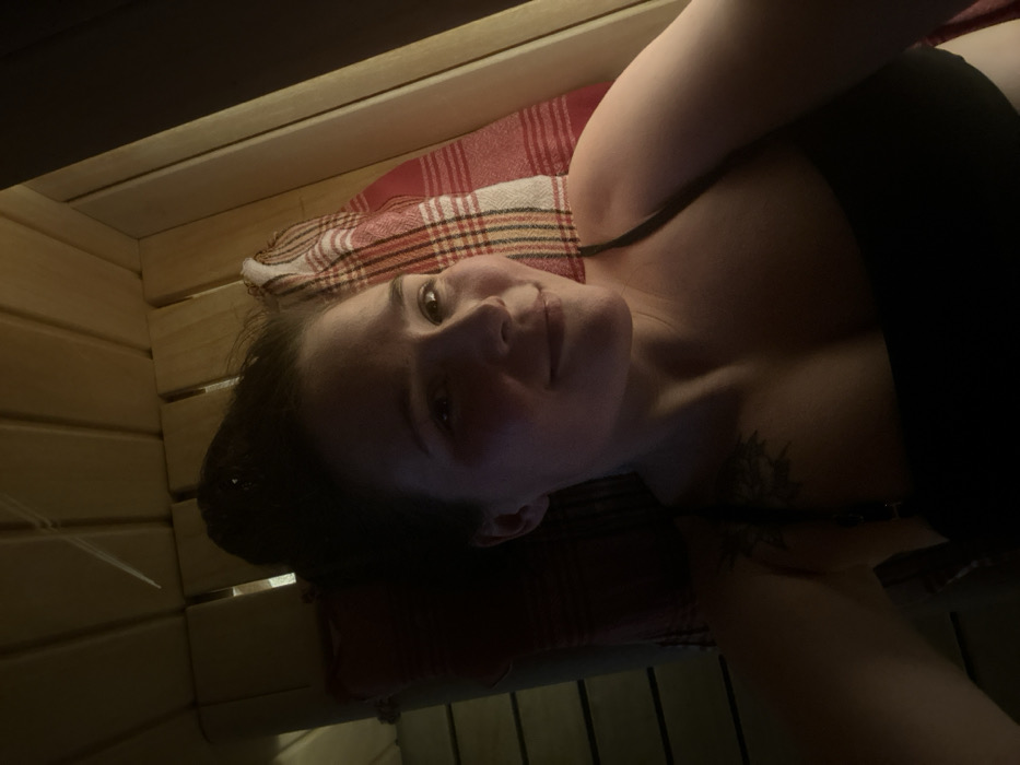 A selfie captures a young adult female reclining on a wooden slatted bench within a dimly lit, wood-paneled room, consistent with a sauna environment. Her dark hair is gathered into a high bun. She is wearing a black strappy top, and a dark tattoo is visible on her left shoulder. Her head rests on a red and white plaid towel. She exhibits a relaxed expression with a slight smile, looking directly at the camera. Warm, soft light illuminates the scene primarily from the right side of the frame, casting shadows that highlight the wooden structure and portions of her face. The specific location within Bartula, Montenegro, cannot be determined from the visual information presented in the image.