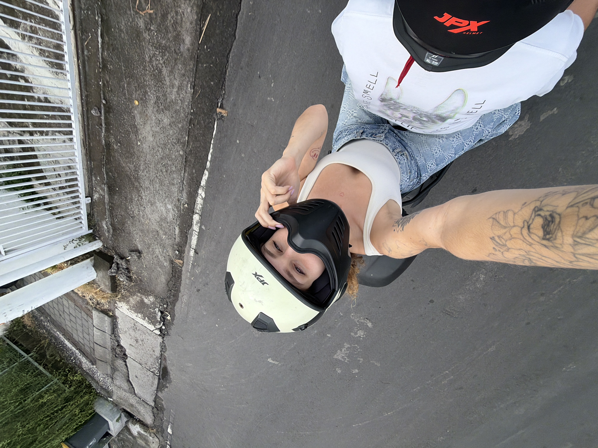 In North Kuta, Indonesia, a person wearing a light-colored JPX helmet with a black face mask, a white tank top, and distressed denim jeans is captured from a high-angle perspective while riding on the back of a motorcycle. The person's left hand is adjusting the helmet's face mask. Another person's arm, tattooed with floral designs, is visible in the foreground, extending to the left and right. A white t-shirt with a graphic print and red accents is worn by the driver of the motorcycle, with the text "SMELL" partially visible. The motorcycle is traveling on an asphalt road with a white line marking its edge. To the left of the road, a concrete sidewalk is broken and uneven, with a grey trash bin situated near a dense green hedge. A white metal fence runs along the right side of the road. The overall scene suggests an outdoor journey on a paved surface within a semi-urban or residential area.
