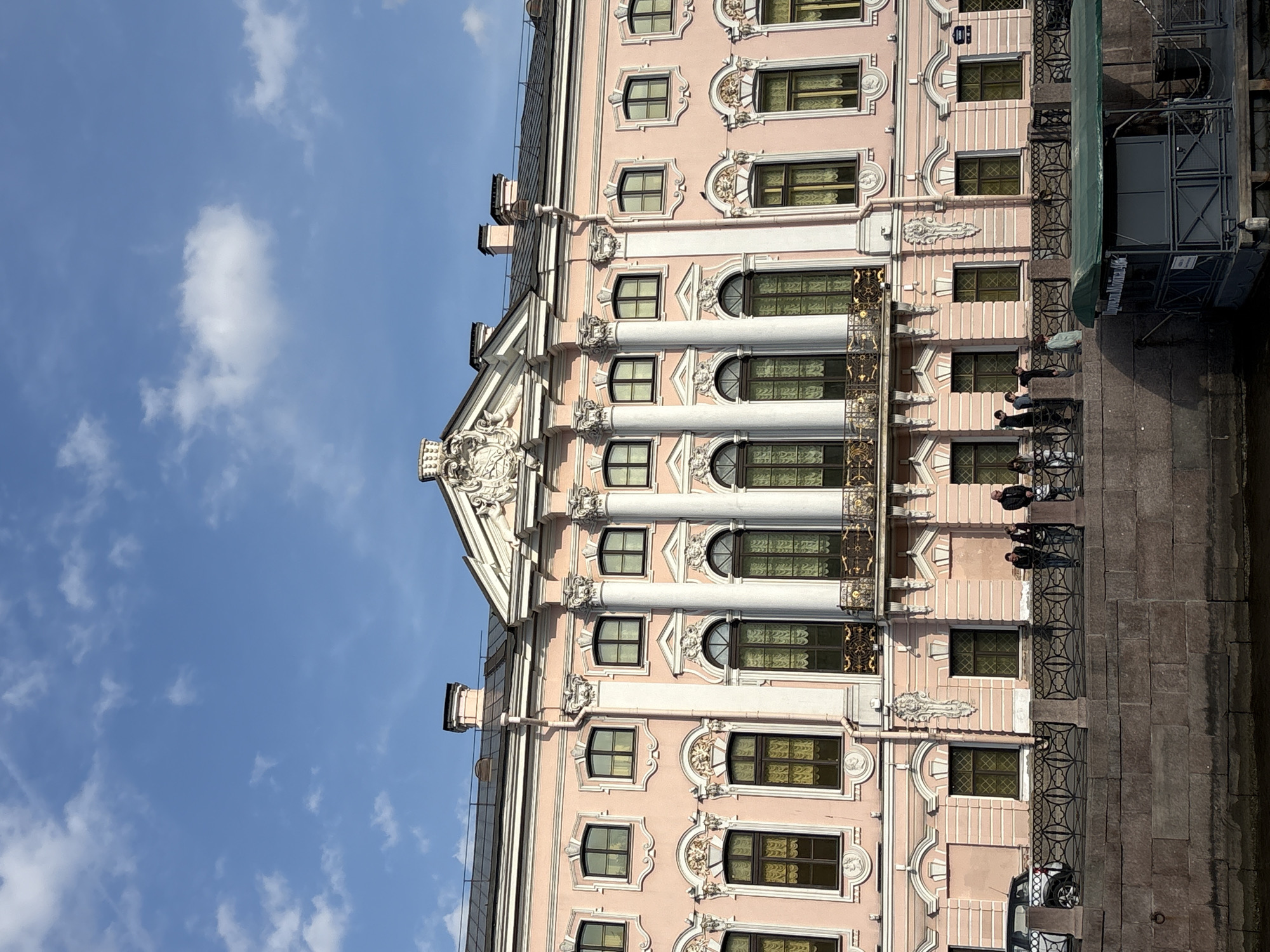 The image shows a side view of a building in Saint Petersburg, Russia. The building is pink with white architectural details, including columns, window frames, and decorative molding. The building has multiple stories and numerous windows. A balcony runs along the building's facade, and several people are standing on it. A stone walkway runs along the side of the building. The sky is blue with scattered clouds.
