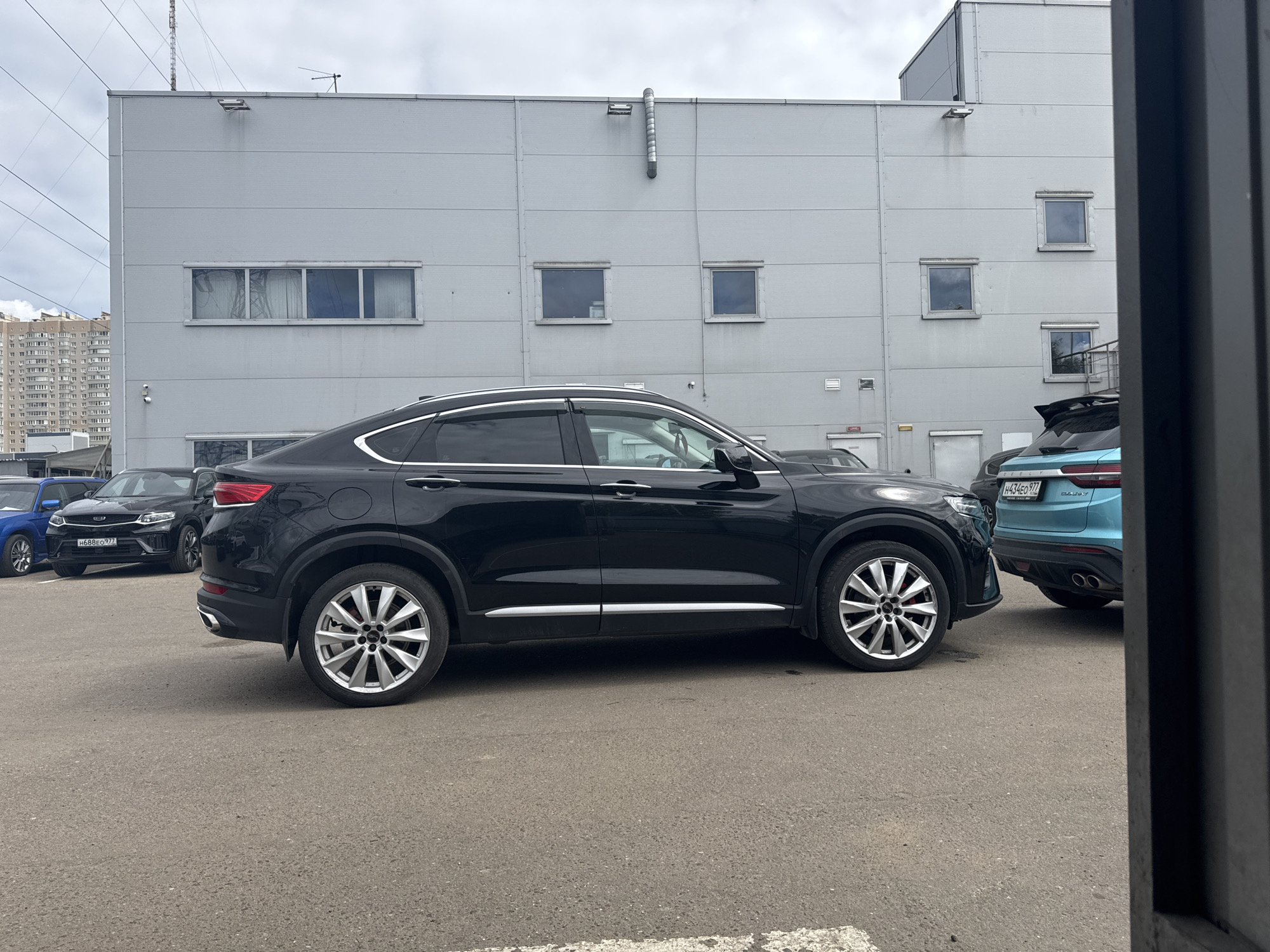 The image depicts a black Geely Tugella coupe SUV parked on an asphalt surface. It is situated in front of a gray, commercial building with a series of square windows on its facade. The background shows a cityscape with apartment buildings, potentially in Balashikha, Russia. To the left of the Geely Tugella is another black car and a blue car partially visible. To the right of the Geely Tugella is a light blue Geely Galaxy, with its license plate partially visible. The composition appears to be taken from a low angle, possibly through a doorway. There are overhead power lines visible in the upper left corner of the frame.