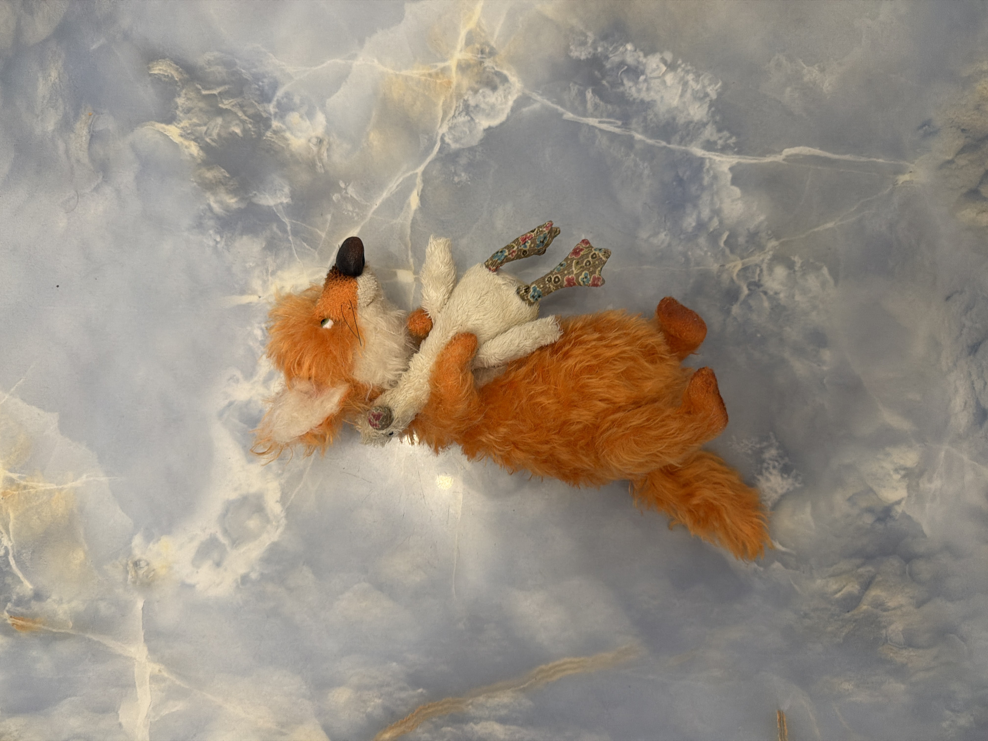 A plush fox toy is held by the arm, with its body angled to the left and its head turned to the right. The fox is predominantly orange with a white chest, muzzle, and ear tips, and a dark brown nose. It appears to be hugging a white plush toy with floral patterned legs. The background is a marbled surface with blue and white veins, possibly a countertop or wall. The image is top-down and well-lit. The location is Unknown, Unknown.