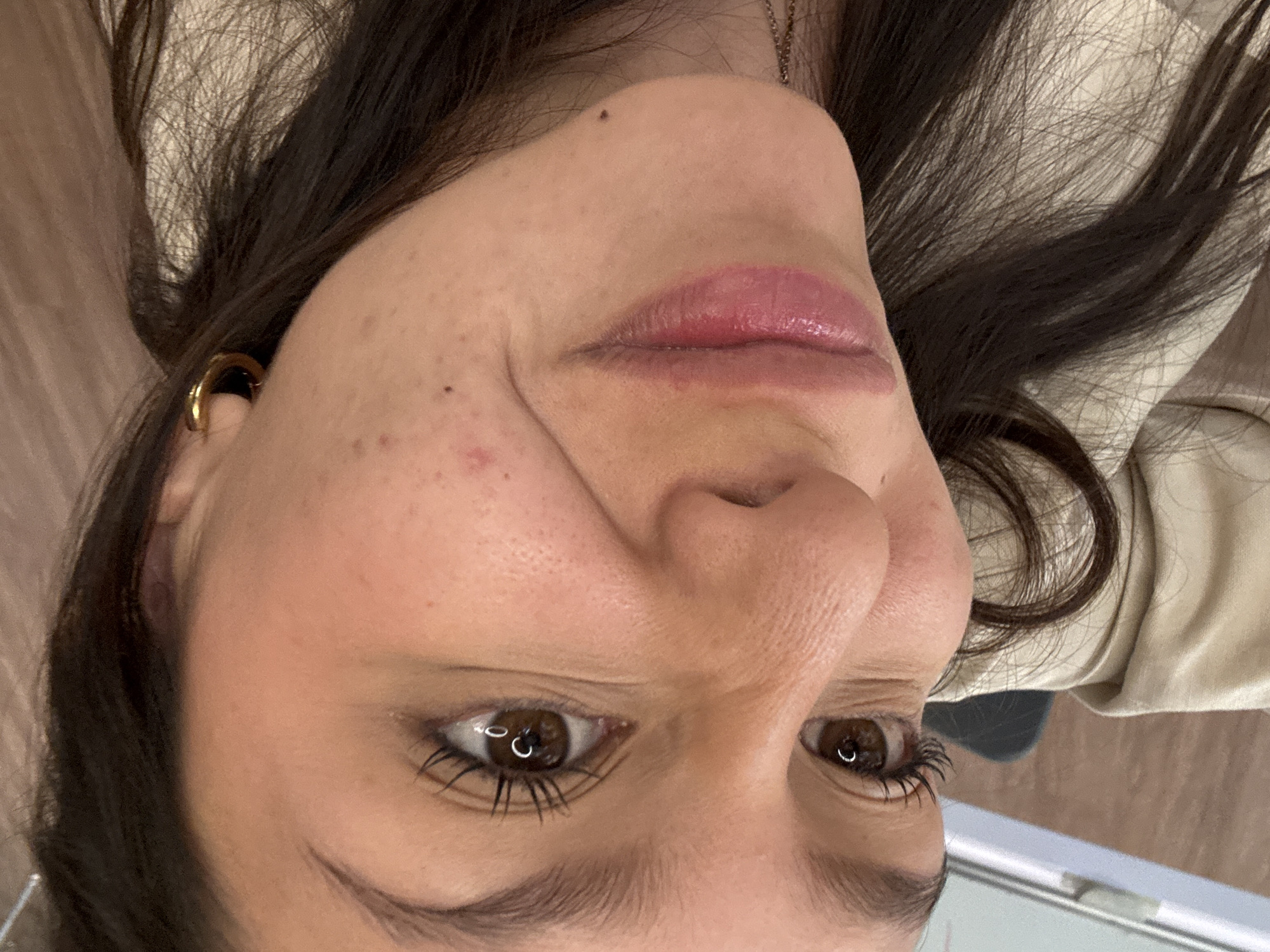A rotated close-up photograph depicts the face of a single female subject in an indoor setting within San Andrés Cholula, Mexico. The image is oriented approximately 90 degrees counter-clockwise, presenting the subject's head tilted towards the left. The subject has long, dark brown hair that frames the face, brown eyes with prominent dark eyelashes, and reflected "C"-shaped ring light sources visible in both pupils. The skin exhibits a tan complexion with visible pores, a small reddish blemish on the forehead, and another below the right eye, along with a small dark mole near the left corner of the mouth. The lips are closed and adorned with a glossy pink or mauve product, with the corners slightly turned downwards, indicating a subtle pout. A gold-colored hoop earring is visible in the subject's right ear, and a thin gold-colored chain necklace is partially visible around the neck. The subject wears a light-toned garment, likely a shirt or jacket, visible around the neck and shoulders. The background is out of focus, showing indistinct indoor elements, including a blurred light-colored vertical structure on the far left.