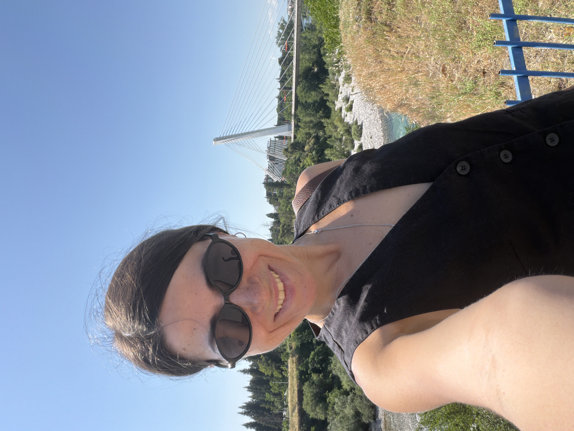 A woman with dark hair and sunglasses takes a selfie outdoors. She is wearing a black vest with buttons and a silver necklace. Behind her, a modern white suspension bridge spans a river, with lush green trees and buildings visible in the background. The sky is clear blue, and the sun is shining. This scene is located in Podgorica, Montenegro, likely near the Millennium Bridge.