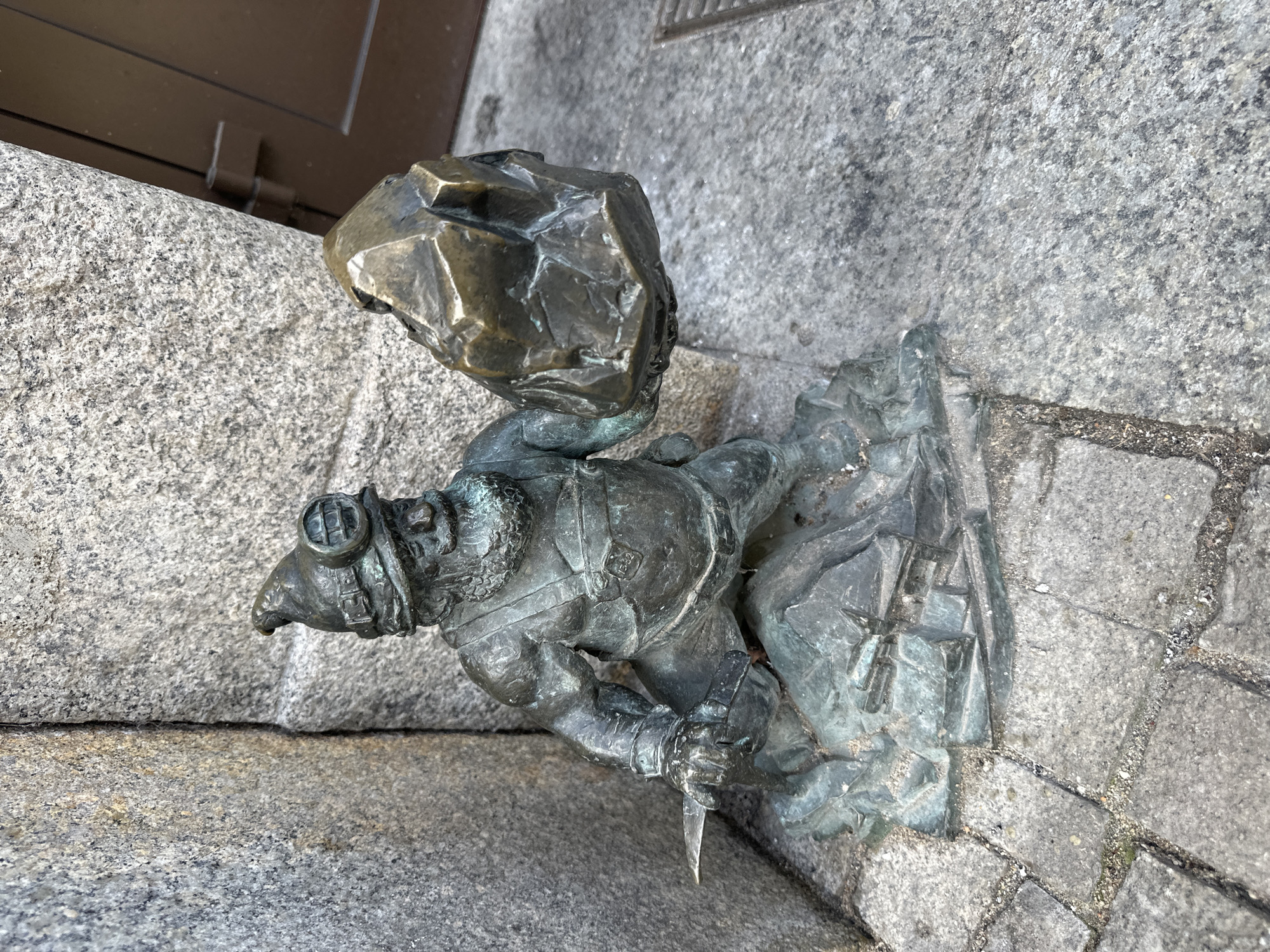 A bronze dwarf statue is positioned on a gray stone surface. The dwarf, wearing mining gear, including goggles on his head and a pickaxe in his hand, carries a large rock on his shoulders. The statue is placed on what appears to be a paved area or building facade in the city of Wrocław, Poland. The stone surface features joints and variations in color and texture. There are hints of building structure in the image background.