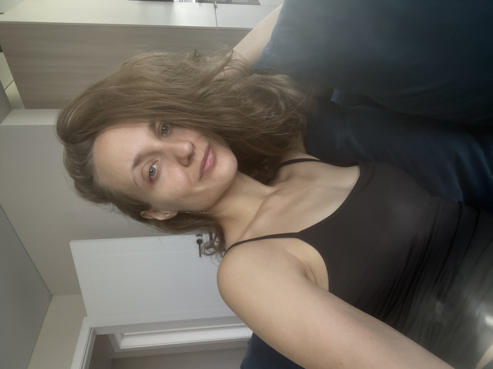 A woman with medium-length brown hair is lying down on a blue couch, looking at the camera. She is wearing a black tank top. The background shows a white door and a light-colored wall. There are no visible urban elements to suggest a specific city location.