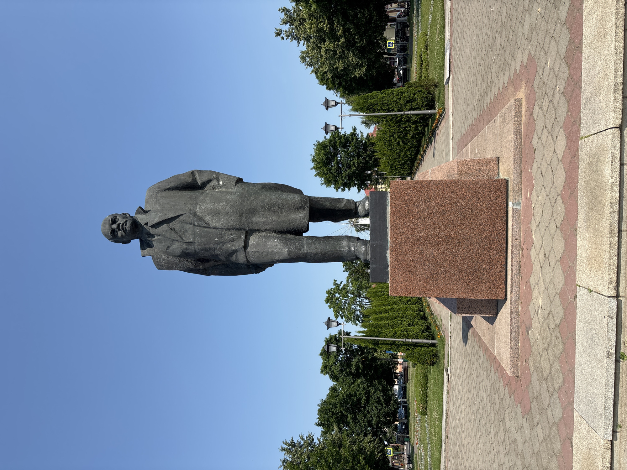 An outdoor, full-shot, eye-level, vertical photograph captures a statue of Vladimir Lenin in Pastavy, Belarus. The statue, rendered in dark metal, depicts Lenin standing with his hands in the pockets of his long coat. He is positioned on a multi-tiered pedestal; the visible portions of the pedestal are made of reddish-brown granite and a darker grey stone base. The statue is centrally located in a paved plaza with a distinctive pattern of grey and reddish-pink interlocking pavers. Flanking the statue on either side are manicured green hedges and mature trees. In the background, a clear blue sky dominates the upper portion of the image. A double-headed lamppost stands to the left of the statue, with another lamppost further down the path to the right. Beyond the plaza and greenery, a street with parked cars and buildings is visible. The overall impression is one of a public monument in a well-maintained urban park or square.