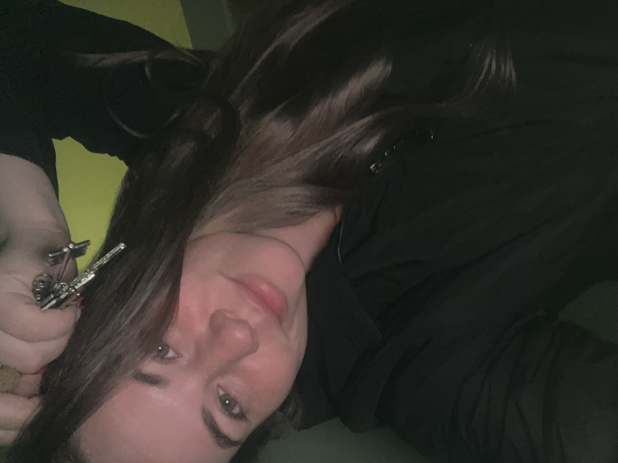A close-up, angled self-portrait depicts a young adult female in the city of Chaikovsky, Russia. The individual possesses long, dark brown hair and light-colored eyes, and is looking directly at the camera with a slight smile. She wears a dark, presumably black, zip-up hoodie or jacket. Her right hand is raised, holding a set of metallic keys on a ring, positioned against the top of her head. The background is dimly lit and indistinct, revealing muted green and yellow hues, consistent with an indoor setting.