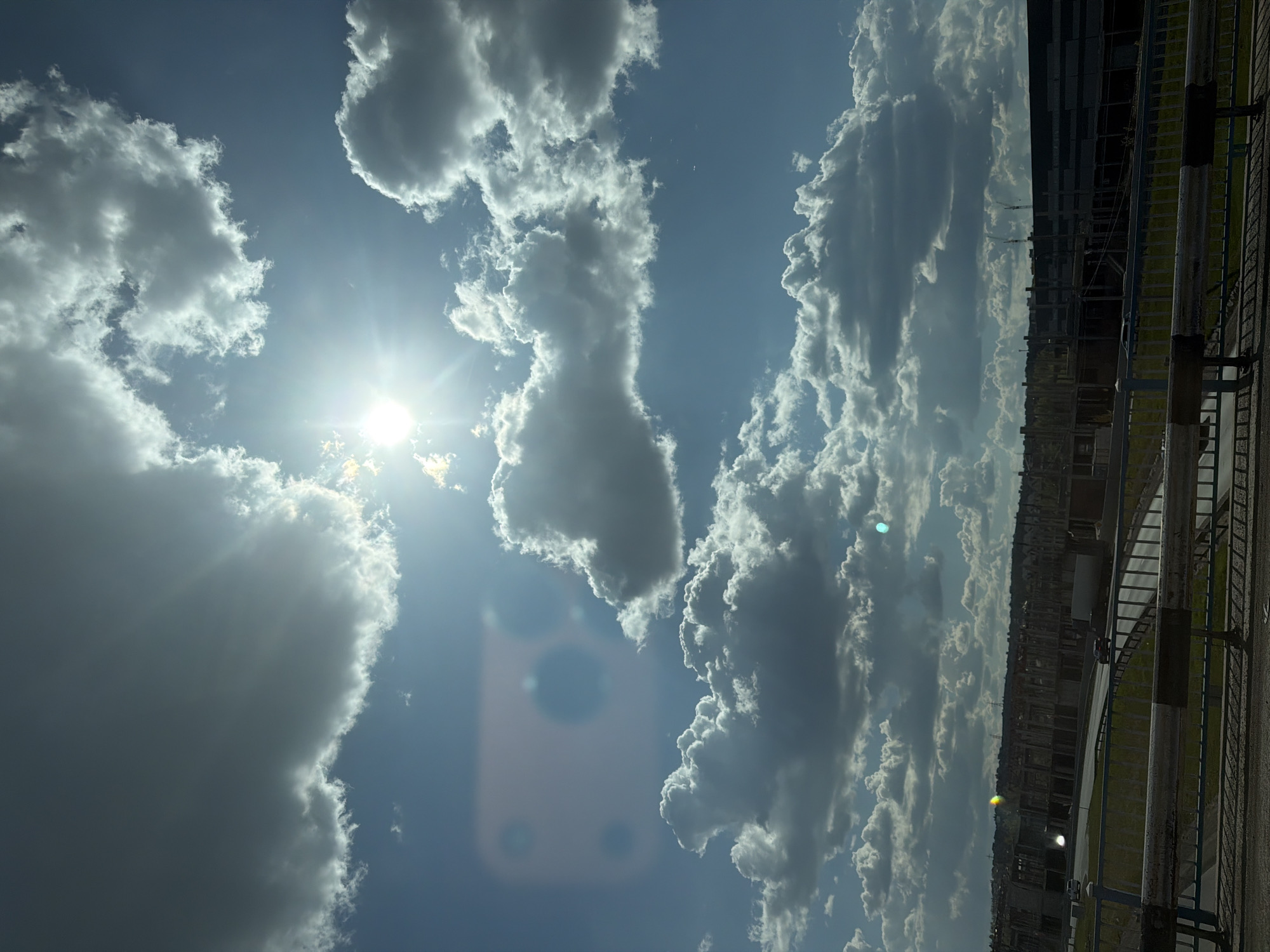 The image shows a daytime scene with a bright sun shining amidst white and gray clouds in a blue sky. In the foreground, a metal railing and part of a road or pavement are visible. Beyond the railing, there's a blurry cityscape with buildings under construction or demolition. The overall location appears to be an urban area on a sunny day. The location is in the city of Unknown, Unknown.