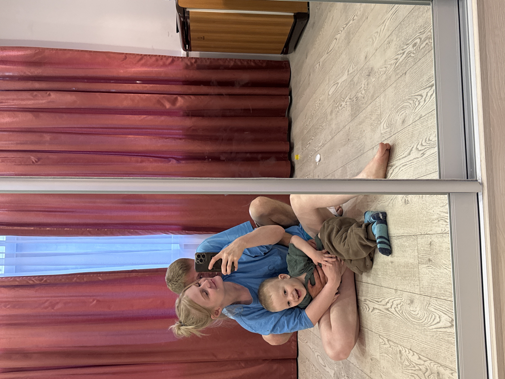 A mirror image shows a family taking a selfie in a room. A woman and a man are seated on the floor with a young child between them. The woman is holding the child, who is laughing and looking at the camera. The man is behind the woman, with his arms around her. All three are wearing casual clothes. The room has wooden flooring and red curtains in the background. The image is taken in Saint Petersburg, Russia.