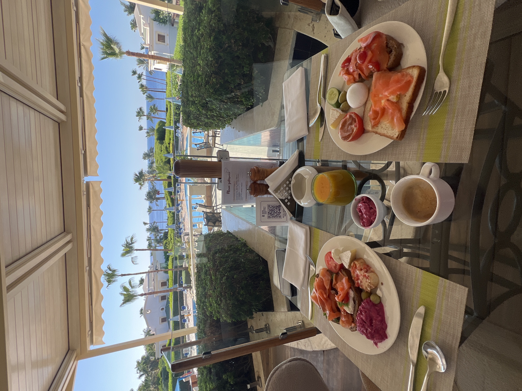The image shows a dining table set for breakfast at a resort in Sharm El Sheikh, Egypt. The table is set with two plates of food, a cup of coffee, a glass of orange juice, and cutlery. The food includes sandwiches with smoked salmon, cheese, and vegetables. In the background, a swimming pool and palm trees are visible, along with a view of the sea. The overall scene depicts a leisurely breakfast enjoyed in a tropical setting.