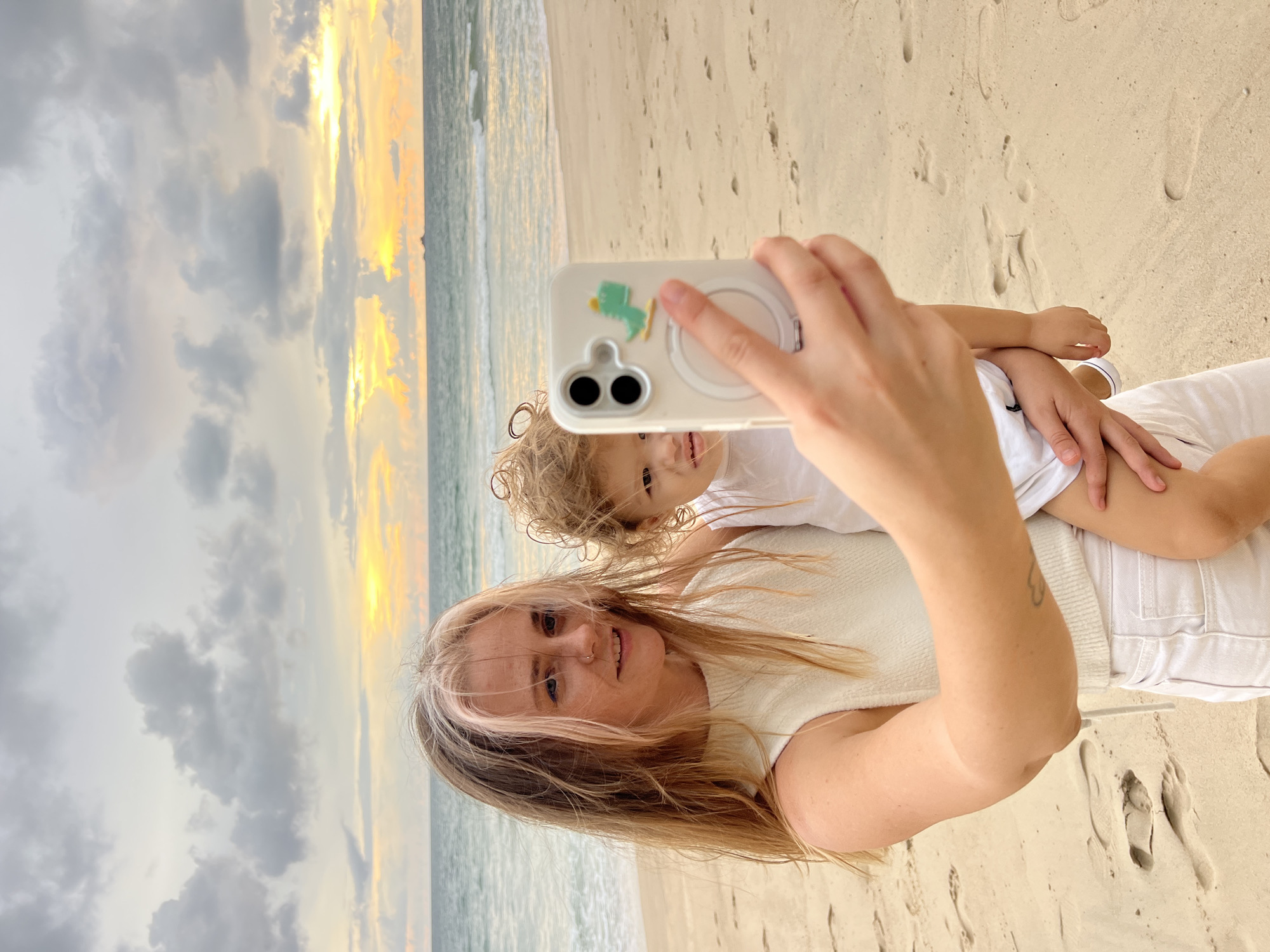 A woman and a toddler are taking a selfie on a beach at dusk. The woman is holding the toddler in her arms and taking the photo with a smartphone. She has blonde hair with pink highlights and is wearing a white sleeveless top and white pants. The toddler has curly brown hair and is wearing a white shirt. The background shows the ocean and a cloudy sky with the sun setting. The sand on the beach is visible, with footprints scattered across it.