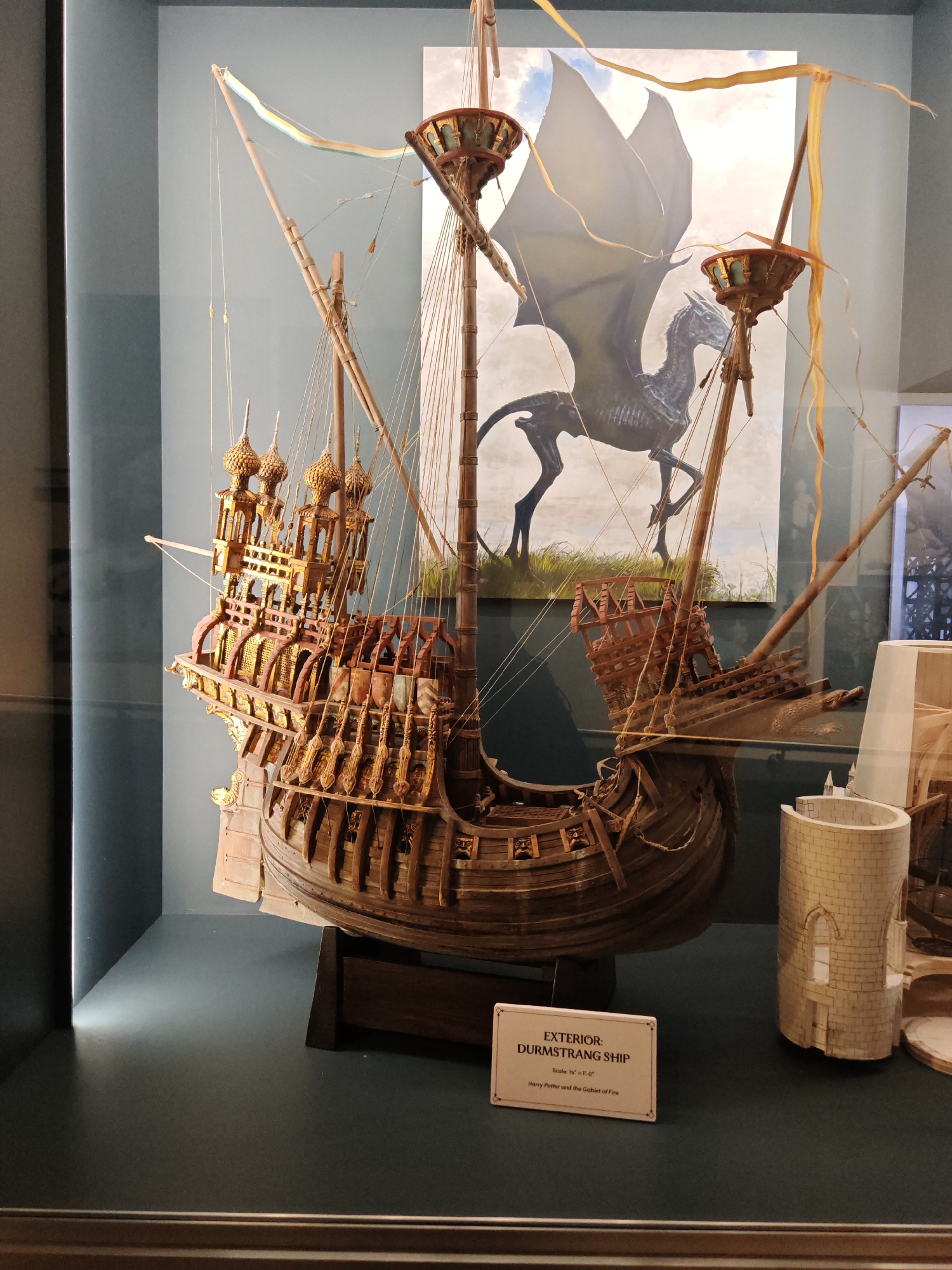 The image is of a model of the Durmstrang ship from the movie "Harry Potter and the Goblet of Fire". The model is housed in a glass display case at Warner Bros. Studio Tour London in Leavesden, Watford, United Kingdom. The model is of a large, ornate wooden ship with multiple masts and sails. The stern of the ship is decorated with intricate carvings and towers. Behind the ship is a backdrop depicting a winged horse-like creature in a field. To the right of the ship is a model of a stone tower. In front of the ship is a small sign that reads "EXTERIOR: DURMSTRANG SHIP" and lists the scale as ½" = 1'-0" and the movie title "Harry Potter and the Goblet of Fire".