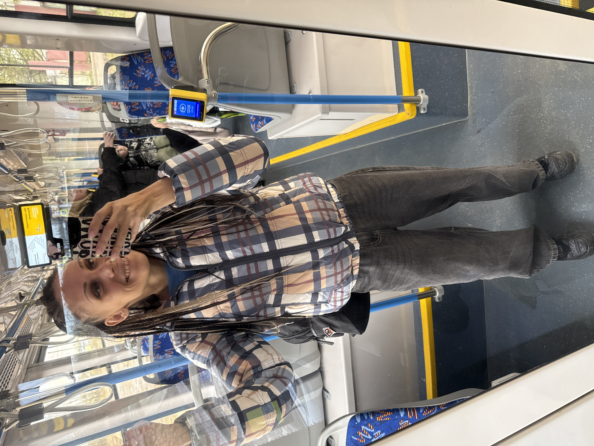 A woman takes a selfie inside a public transport vehicle, likely a bus or tram, based on the interior design. The image is captured from her perspective, showing her reflection in a window or glass partition.
The woman has dark hair styled in dreadlocks or braids, and is smiling directly at the camera (her phone). She is wearing a plaid jacket in shades of brown, beige, blue, and white, over a blue top, and dark grey or black jeans. Her right hand holds the phone, which has a patterned case.
The setting appears to be modern public transportation. The floor is dark grey with a speckled pattern, and there are yellow safety lines marking areas. Blue metal grab poles run horizontally, attached to the ceiling and sidewalls. One such pole has a yellow ticket validator device mounted on it, displaying a blue screen with white icons and text, indicating it's active.
Through the windows, which also reflect parts of the vehicle's interior, glimpses of the outside world can be seen, suggesting it's daytime. Distant buildings and trees are visible. Other passengers, mostly blurry reflections, can be discerned in the background, including one person in a dark jacket. The seats in the background have blue upholstery with a white or light-colored pattern.
Visible text on the ticket validator screen, though small, appears to be in a Cyrillic script, displaying "ПРИЙНЯТО" (Accepted) or similar confirmation, and other interface elements typical for payment or validation. Another yellow sign with Cyrillic text is reflected on the upper left, possibly providing route information or instructions.