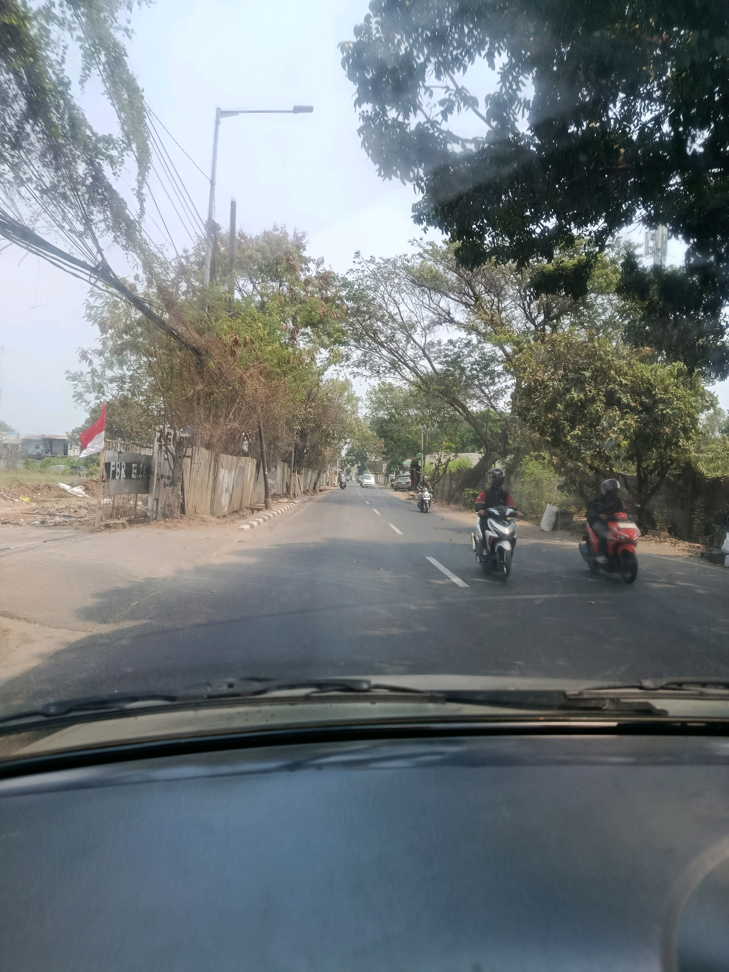 The image captures a view from inside a vehicle, looking out onto a street in East Jakarta, Indonesia. The street is paved and appears to be two lanes wide. Several motorcycles are visible on the road, with two riders in the foreground of the frame. One motorcycle is white and red, and the other is red. Trees line the right side of the road, and a wooden fence is visible behind them. On the left side of the road, there is an undeveloped area with some construction materials and a large red and white flag. A street lamp is also visible on the left side of the image. The sky is bright and hazy, suggesting it is daytime. The overall scene depicts a typical urban streetscape in a tropical climate.