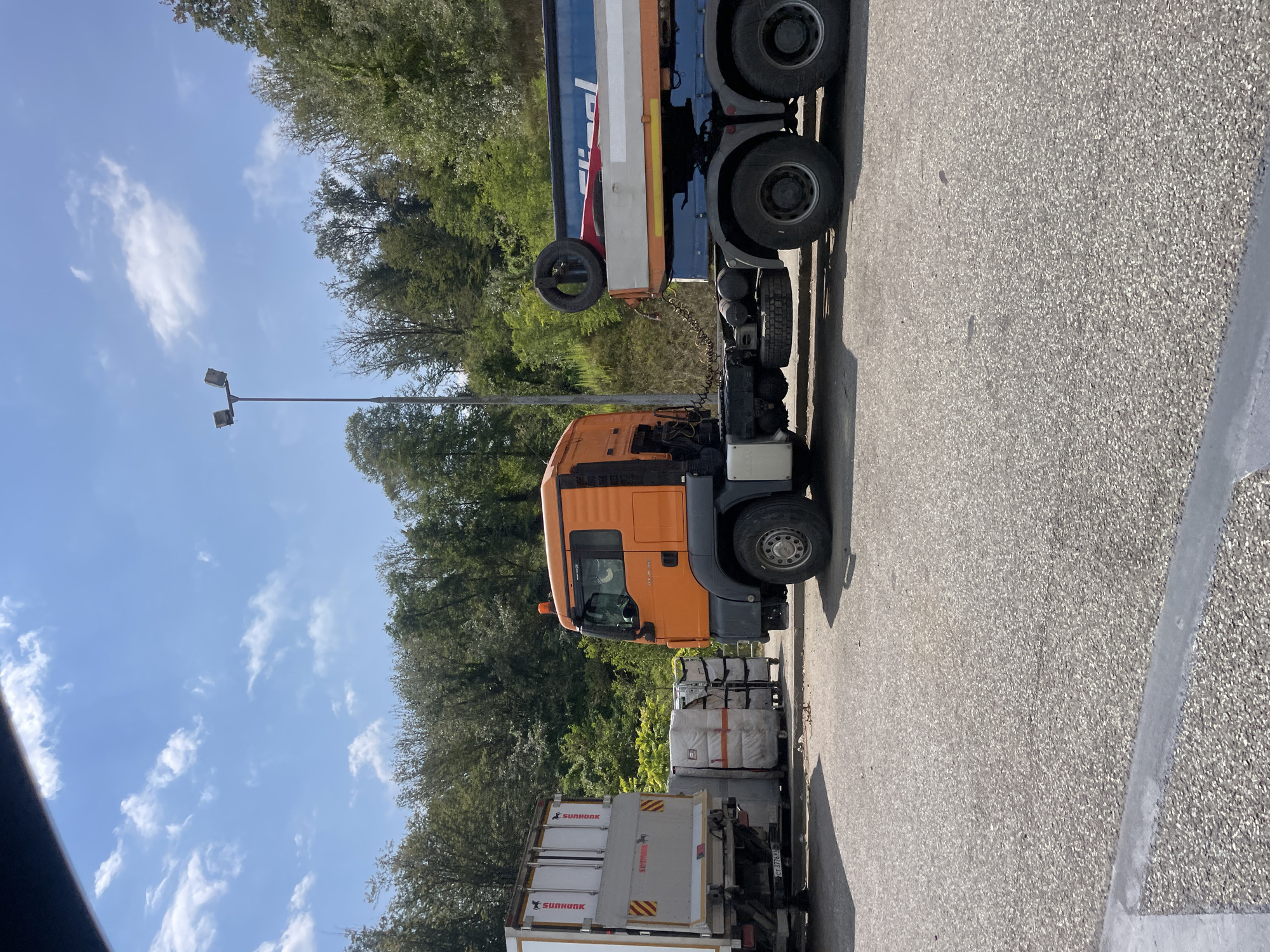 A daytime, slightly low-angle outdoor shot shows two semi-trucks parked on a paved lot next to a wooded hillside. The truck on the left has a white and grey trailer and appears to be loaded with what looks like boxed goods. The truck on the right is an orange tractor pulling a blue and grey trailer with a spare tire mounted on its side. The sky is blue with scattered white clouds. A tall street lamp with two lights is visible to the left of the orange truck. The trees on the hillside are a mix of green and brown, suggesting it may be late spring or early summer. The ground is asphalt with some scattered debris. The scene is calm and still, with no people visible.