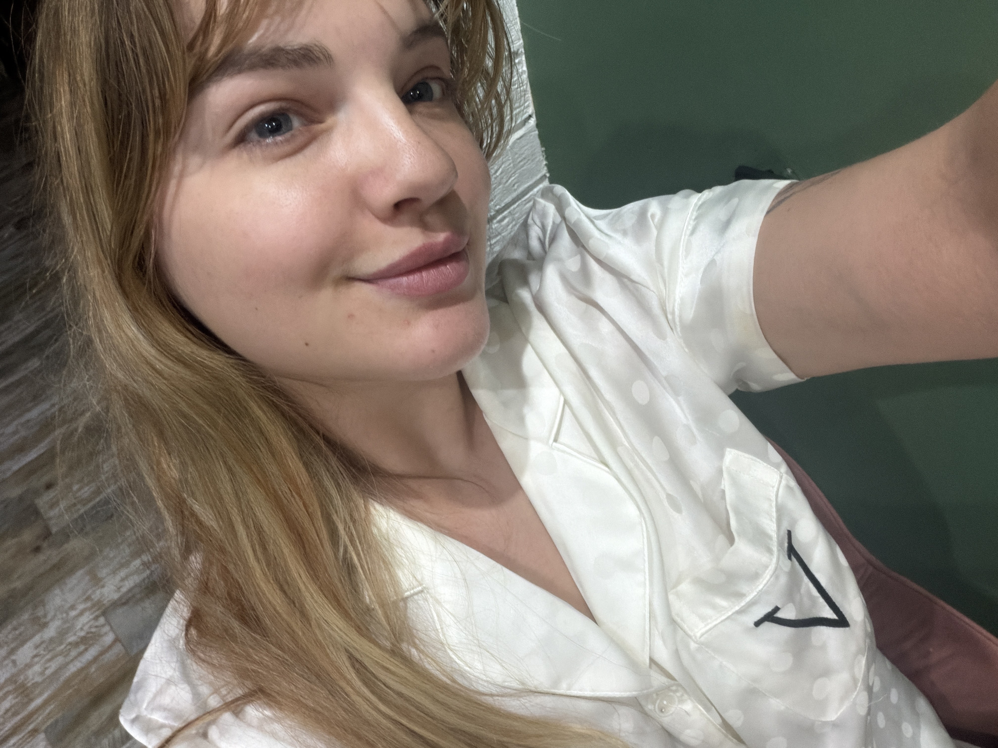 A woman with fair skin, light brown hair, and blue eyes is taking a selfie. She is wearing a white polka-dot pajama top with a black "V" stitched on the pocket. The background appears to be a wall with green paneling and another wall with a brick or wood texture. Location information indicates that the picture was taken in Большая Шильна, Russia.