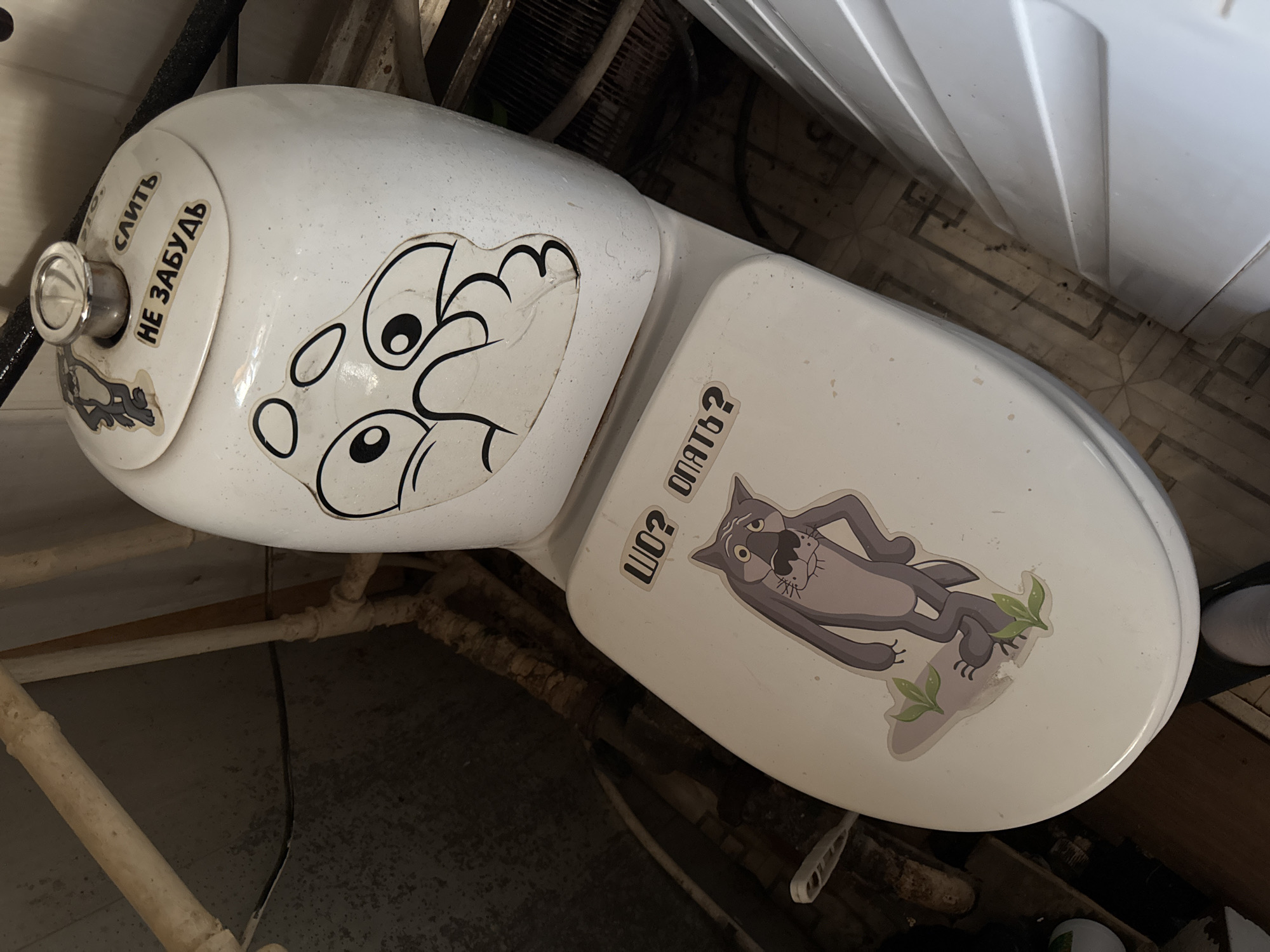 The image shows a white toilet with stickers on the tank and seat. On the toilet tank, there is a sticker of a cartoon character with a concerned expression and text that reads "СЛИТЬ НЕ ЗАБУДЬ" (Don't forget to flush). On the toilet seat, there is a sticker of a cartoon wolf with its hand on its hip and text that reads "ШО? ОПЯТЬ?" (What? Again?). The toilet is in a bathroom with pipes visible in the background. The floor appears to be tiled. The image is taken from a high angle, looking down at the toilet.