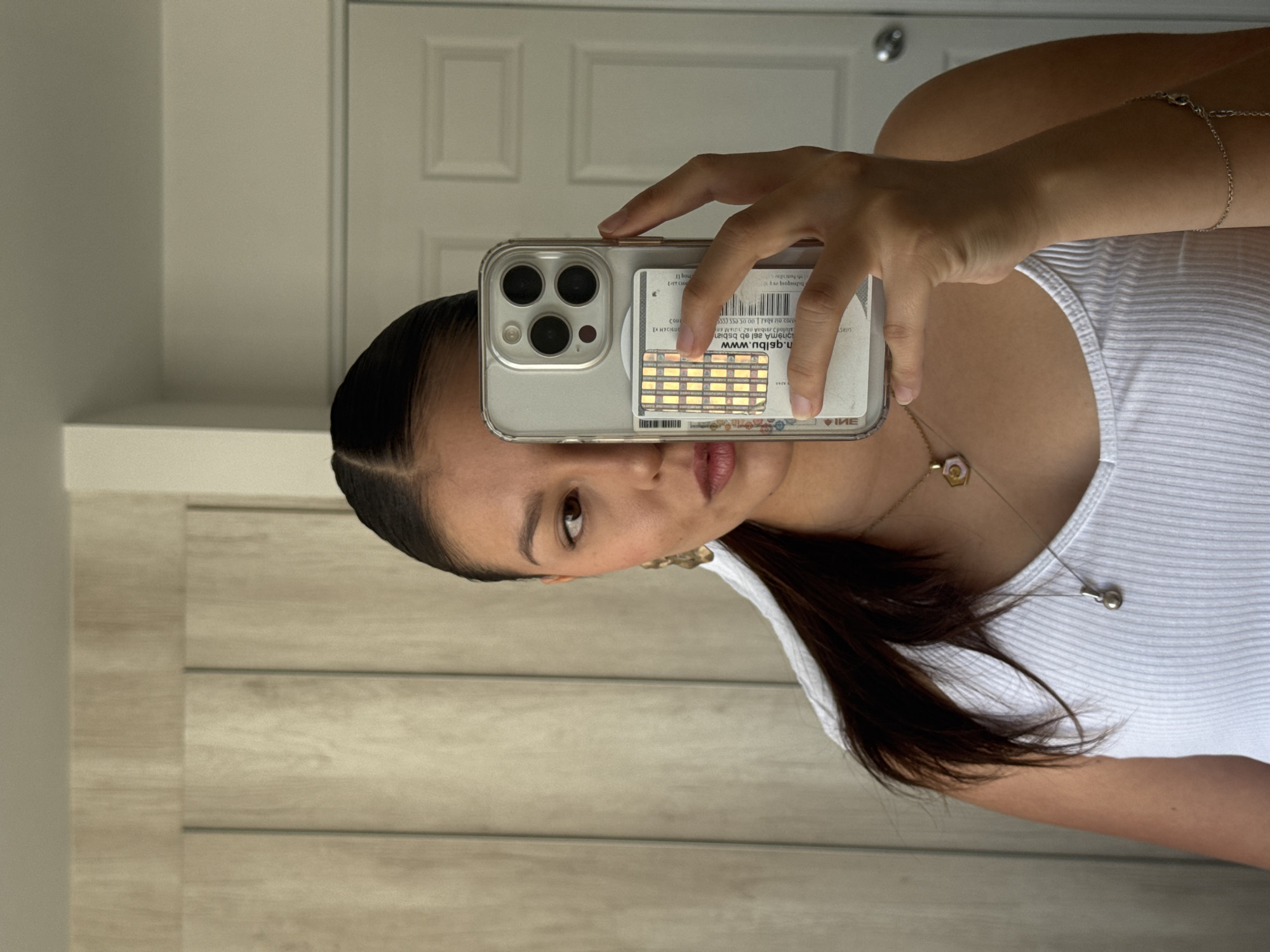 The image shows a woman taking a selfie in what appears to be an indoor environment. She is holding her phone, which has a clear case, up to her face. Behind the phone, in the case, are a credit card and a card with a QR code and website address printed on it. The woman has dark hair pulled back from her face, is wearing a white tank top, and has a bracelet on her wrist, a necklace with a flower pendant, and a second necklace with a small pendant. The background includes a white door with recessed panels and what appears to be a wall with a horizontal wood-like pattern. The location information of San Francisco Ocotlán (Ocotlán), Mexico, is not visible in the image.