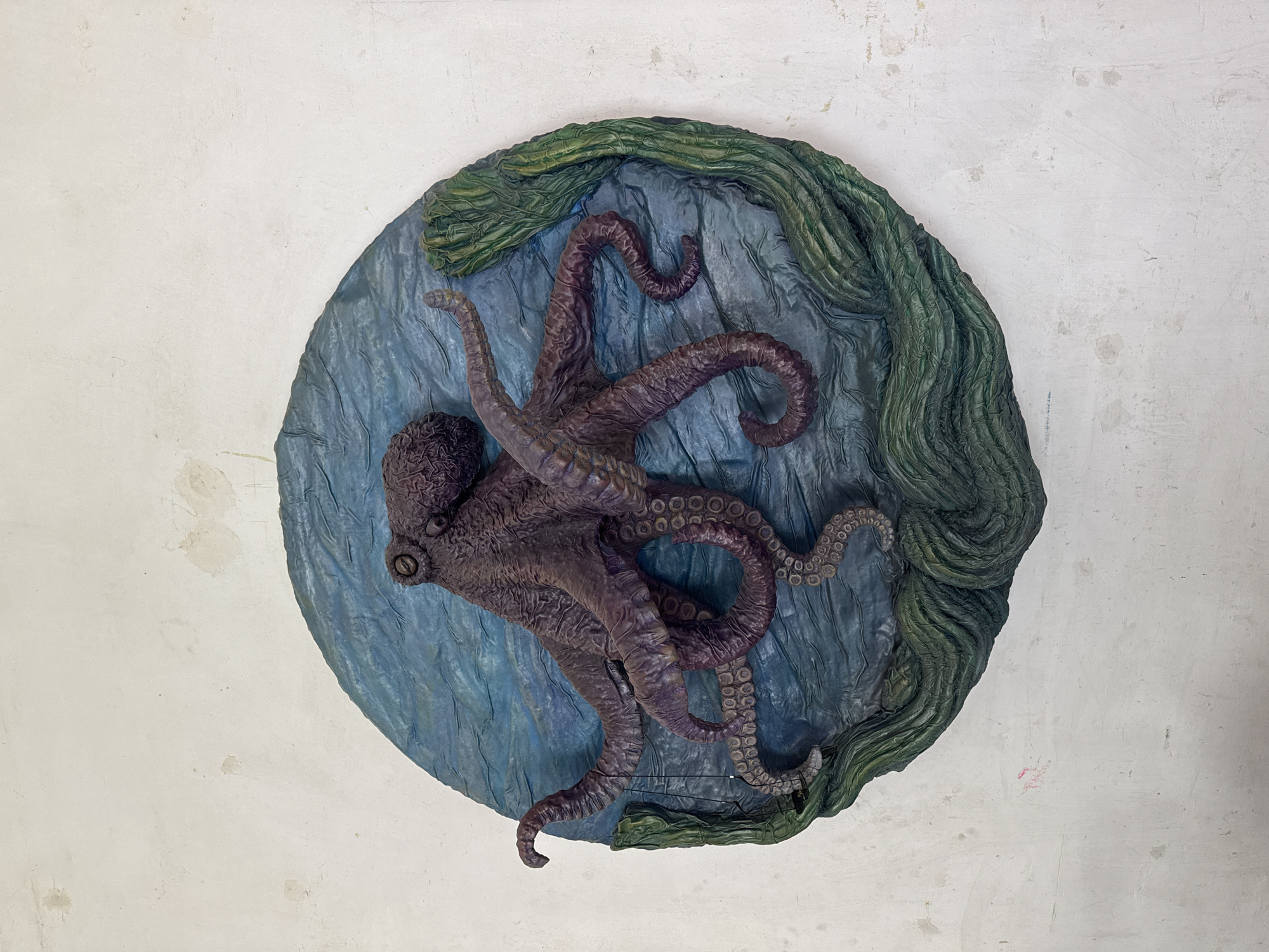 A circular wall plaque depicts a dark purple octopus with eight tentacles spread out on a textured blue background, designed to resemble water. Green, wavy seaweed forms the bottom edge of the plaque. The plaque is affixed to a textured, off-white wall with visible imperfections, including stains and scuffs. The scene is indoors, likely a studio or workshop, and there is no indication of the city of Kolomyia, Ukraine.