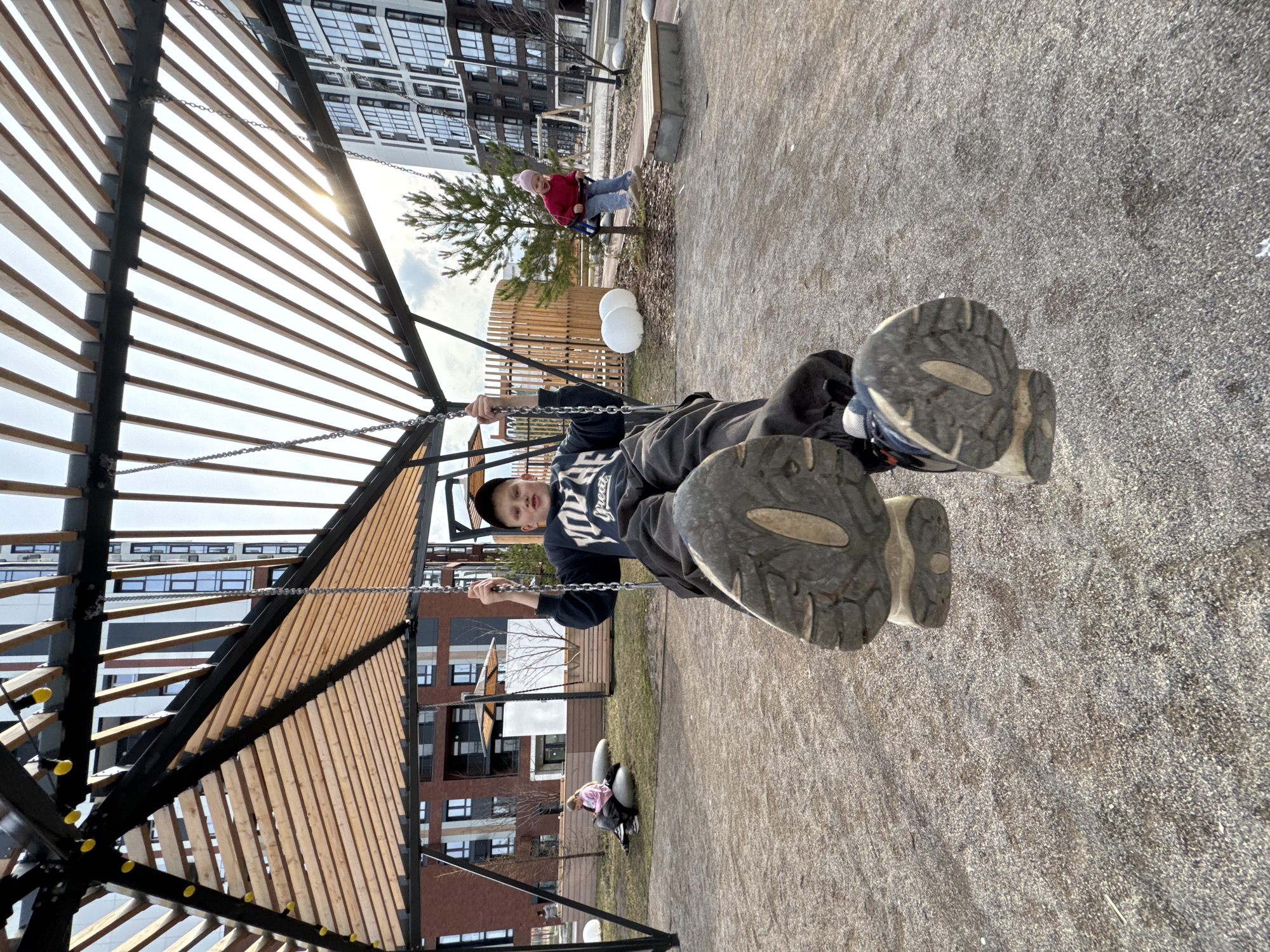 A boy in a black hooded sweatshirt and black cap is riding on a swing set at a playground. The playground features a modern design with wooden slats and metal framework. The ground surface appears to be a compacted granular material. Other playground equipment and children are visible in the background. Apartment buildings are visible in the background. The location is likely in the city of Perm, Russia.