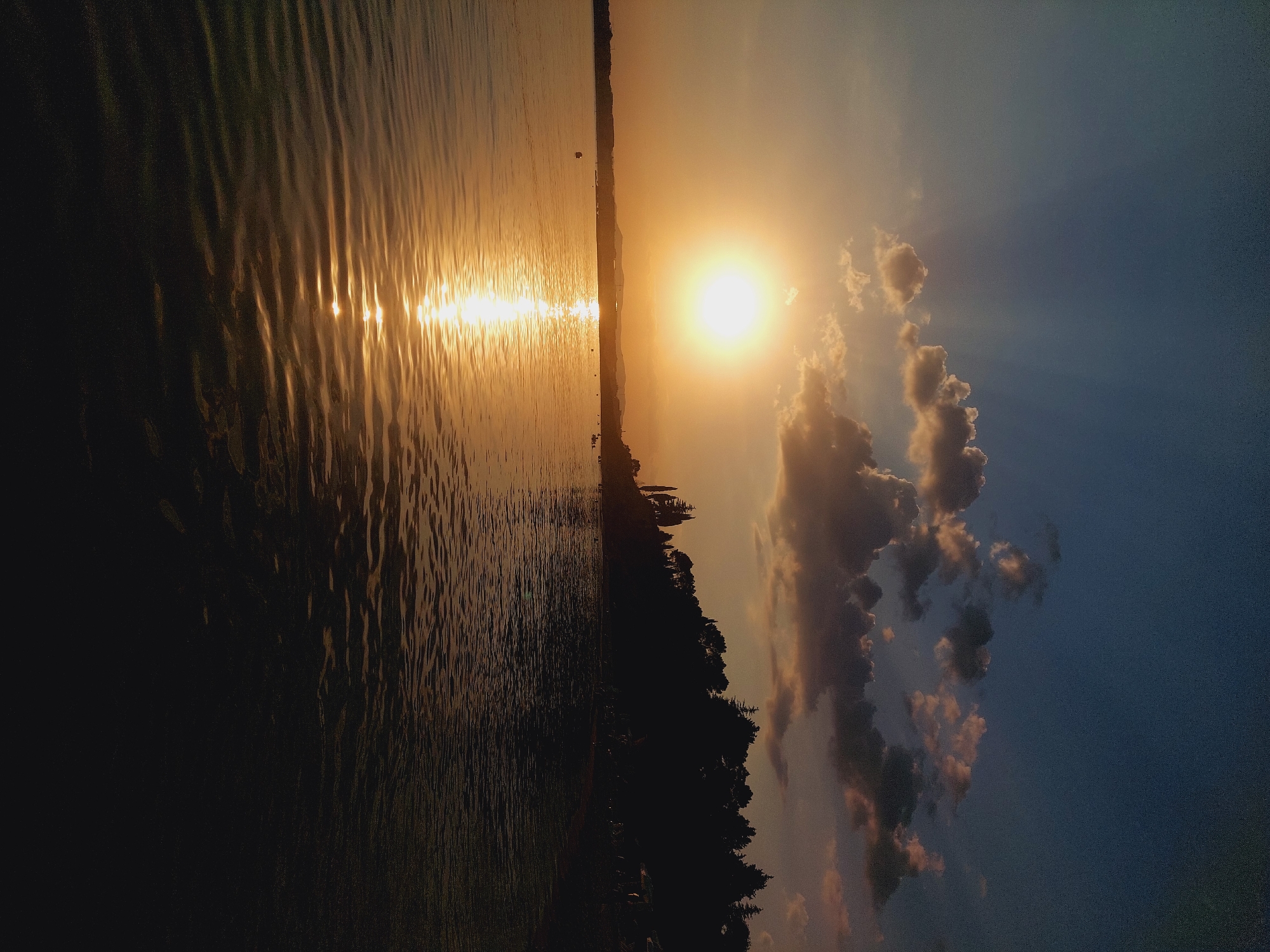 This image depicts a serene sunset over a body of water, likely the sea, with the silhouette of a coastline in the background. The sun, a bright golden orb, is positioned in the upper left quadrant of the frame, casting a shimmering reflection across the rippling water. The sky transitions from a deep blue at the top to lighter, warmer hues near the horizon, punctuated by scattered, illuminated clouds. The coastline is densely forested with trees, creating a dark, jagged silhouette against the fading light. A few human figures are visible as small, indistinct shapes along the shoreline and in the water, suggesting a beach or recreational area. The overall mood is tranquil and picturesque. Without further context or specific landmarks visible, it is not possible to definitively identify the location as Nesebar, Bulgaria.