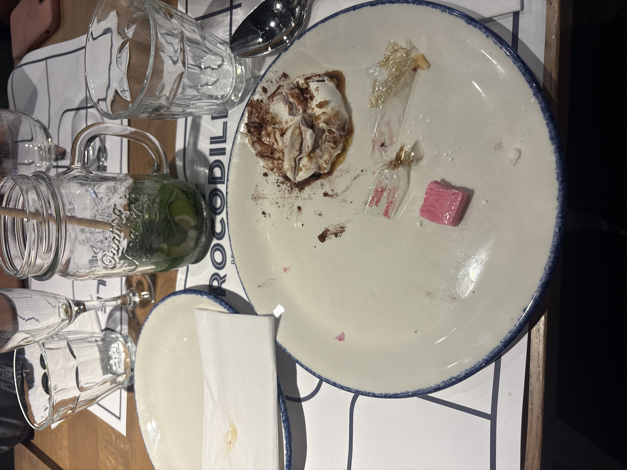 The image displays a cluttered tabletop scene at what appears to be a restaurant or bar, likely after a meal. A large, white ceramic plate with a distinctive blue rim occupies the center, containing the remnants of a dessert – a partially consumed, cream-based dish with chocolate powder and sauce stains. Beside the dessert, two crumpled clear plastic candy wrappers and a single, rectangular pink candy (possibly chewing gum) are scattered on the plate.
A variety of glassware surrounds the plate: a large, empty, clear glass mug with a handle; a clear glass mason jar mug with a handle, holding a green beverage with mint and lime, suggesting a mojito or similar cocktail, complete with a straw; an empty wine glass; and two other empty clear drinking tumblers. A silver spoon rests near the plate, and a stack of white paper napkins is placed on a smaller, empty white plate with a matching blue rim. The table surface is wooden, and a white placemat with a grid pattern is visible underneath, featuring partial text.
The scene indicates a recently concluded casual dining or social event. The visible text on the placemat clearly reads "CROCODIL", strongly suggesting the restaurant chain "Crocodile," which is present in France and aligns with the provided location context of Tourcoing. Partially visible text on one of the candy wrappers includes "CONFISERIE," a French term for confectionery, further supporting a French setting. The table shows signs of recent use, with crumbs and minor spills.