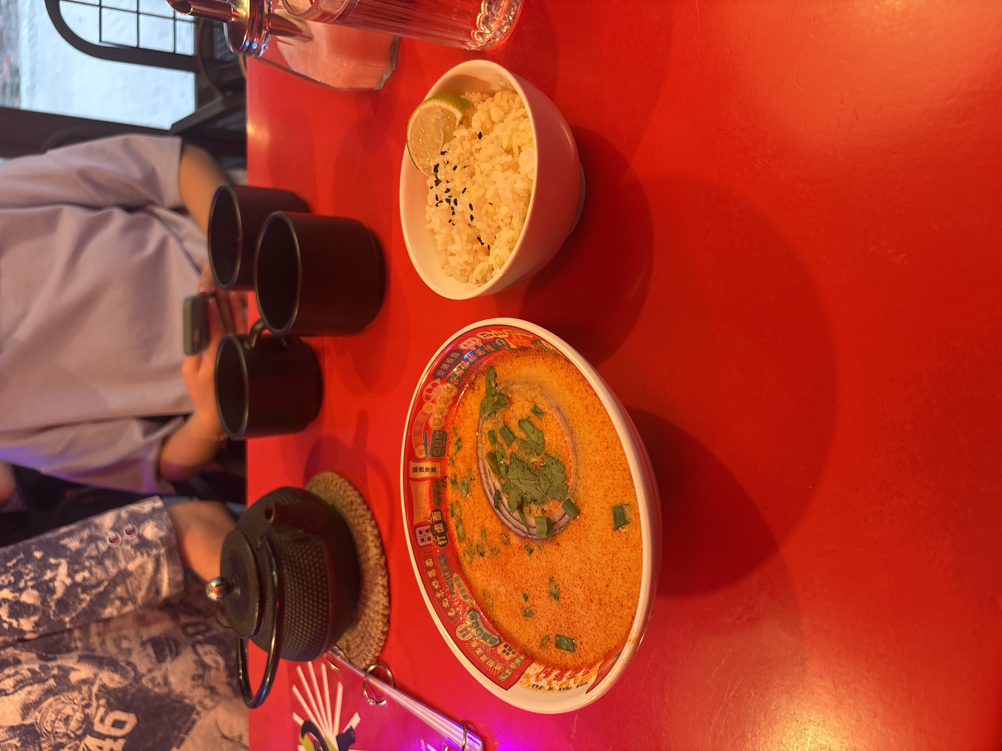 The image depicts a dining table scene, likely in a restaurant in Perm, Russia, as suggested by the presence of dishes and the "MIAN JU DAO" text which translates to "Noodle House" and the restaurant name in the background. The table, covered in a bright red surface, displays an array of items indicative of a meal: a bowl of rice garnished with black sesame seeds and a slice of lime, a bowl of what appears to be soup or noodles, and several dark-colored cups alongside a teapot on a straw placemat. Two individuals are partially visible, one wearing a light purple shirt and the other wearing a blue-grey t-shirt. The scene suggests a meal being enjoyed, possibly at a restaurant specializing in Asian or specifically Chinese cuisine. A clear glass bottle is also present, and the background is slightly blurred.