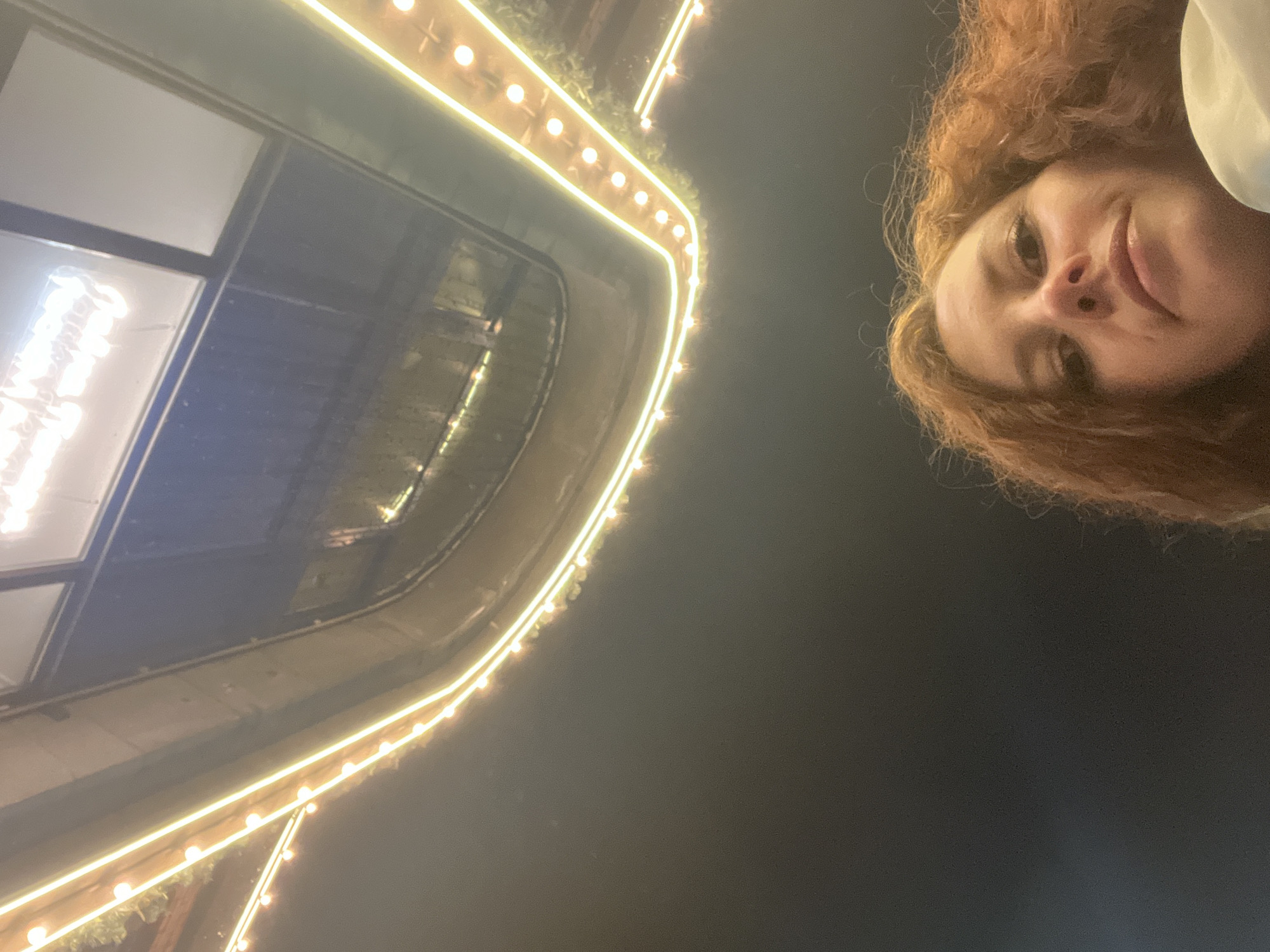 A low-angle shot depicts a female with red, curly hair and fair skin, positioned on the upper right of the frame. She is smiling slightly. In the background, on the left side, there is a building facade with large windows outlined by string lights. The time of day appears to be evening or night. The location is potentially Saint Petersburg, Russia, given the architectural style.