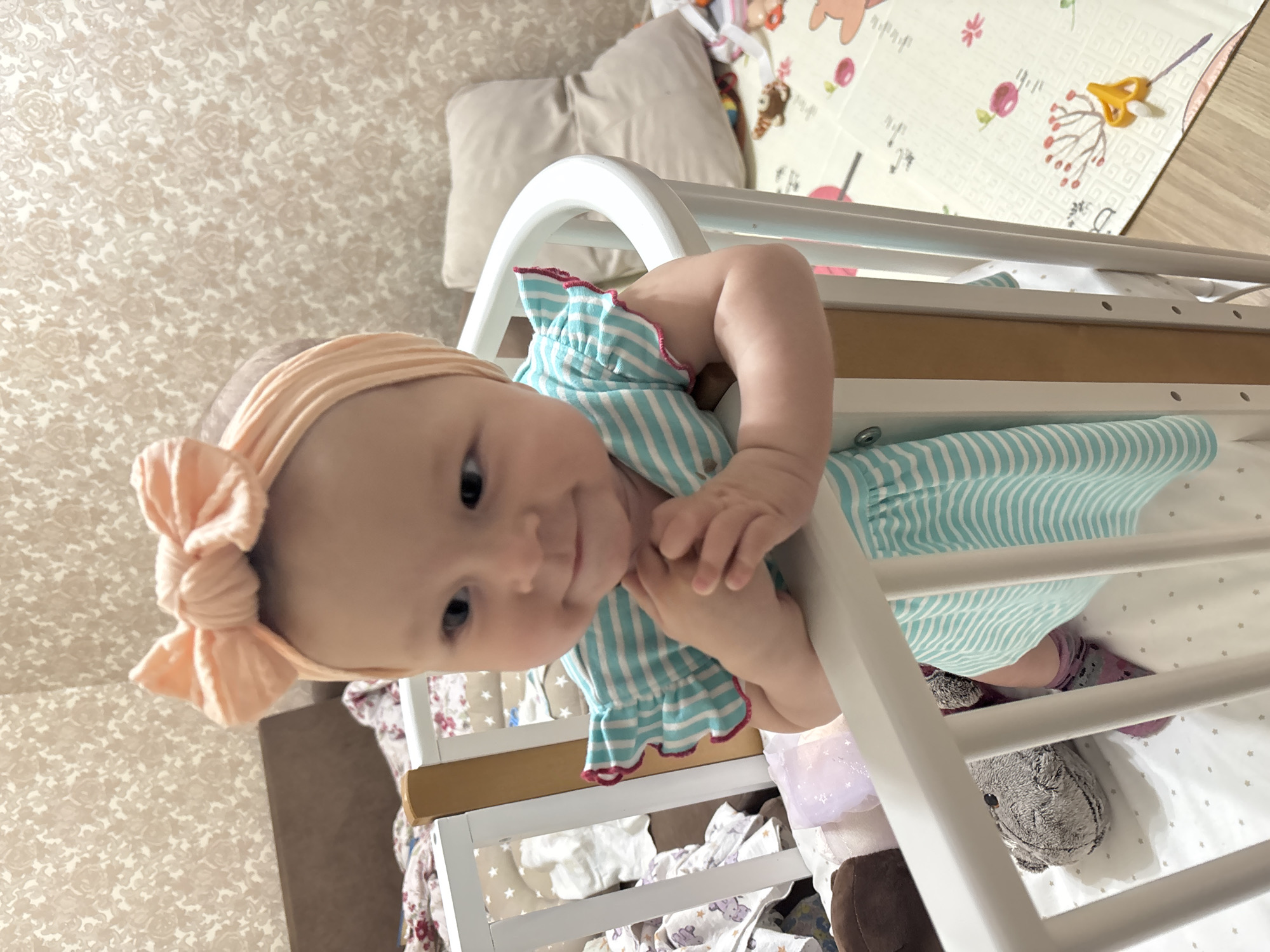 A baby wearing a peach-colored headband with a bow and a blue and white striped dress with pink trim is positioned in a white crib. The baby is holding onto the crib's railing and looking at the camera. Inside the crib, there are toys and blankets. In the background, there's a pillow, a patterned play mat, and a wallpapered wall. The location is likely inside a private home, not specifically identifiable as Sochi, Russia.
