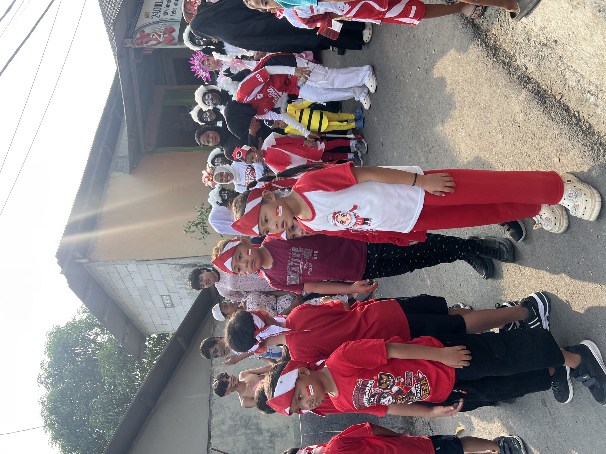 In Klapanunggal, Indonesia, a group of children and adults are gathered outdoors, possibly for a parade or celebration. Many of the children are dressed in red and white attire, with some sporting face paint. One child wears a bumblebee costume. In the background, a building with a corrugated metal roof is visible. The ground appears to be asphalt. A sign with Indonesian text is affixed to the building. The scene suggests a festive and communal gathering, possibly related to national holidays or cultural events.