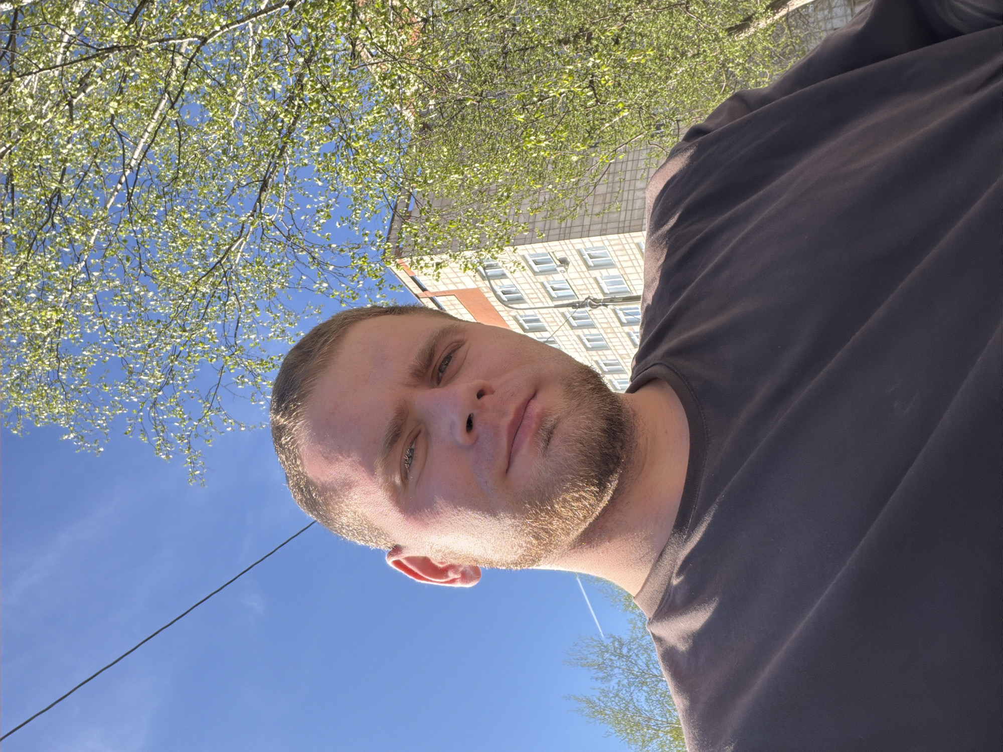 A man with fair skin, short blonde hair, and a beard is the central subject, captured from a low-angle, upward perspective. He is wearing a dark t-shirt. Behind him is a clear blue sky and some tree branches with green leaves. In the background, the upper section of a light-colored brick building with many windows is visible. The photo appears to be taken in Chaikovsky, Russia.