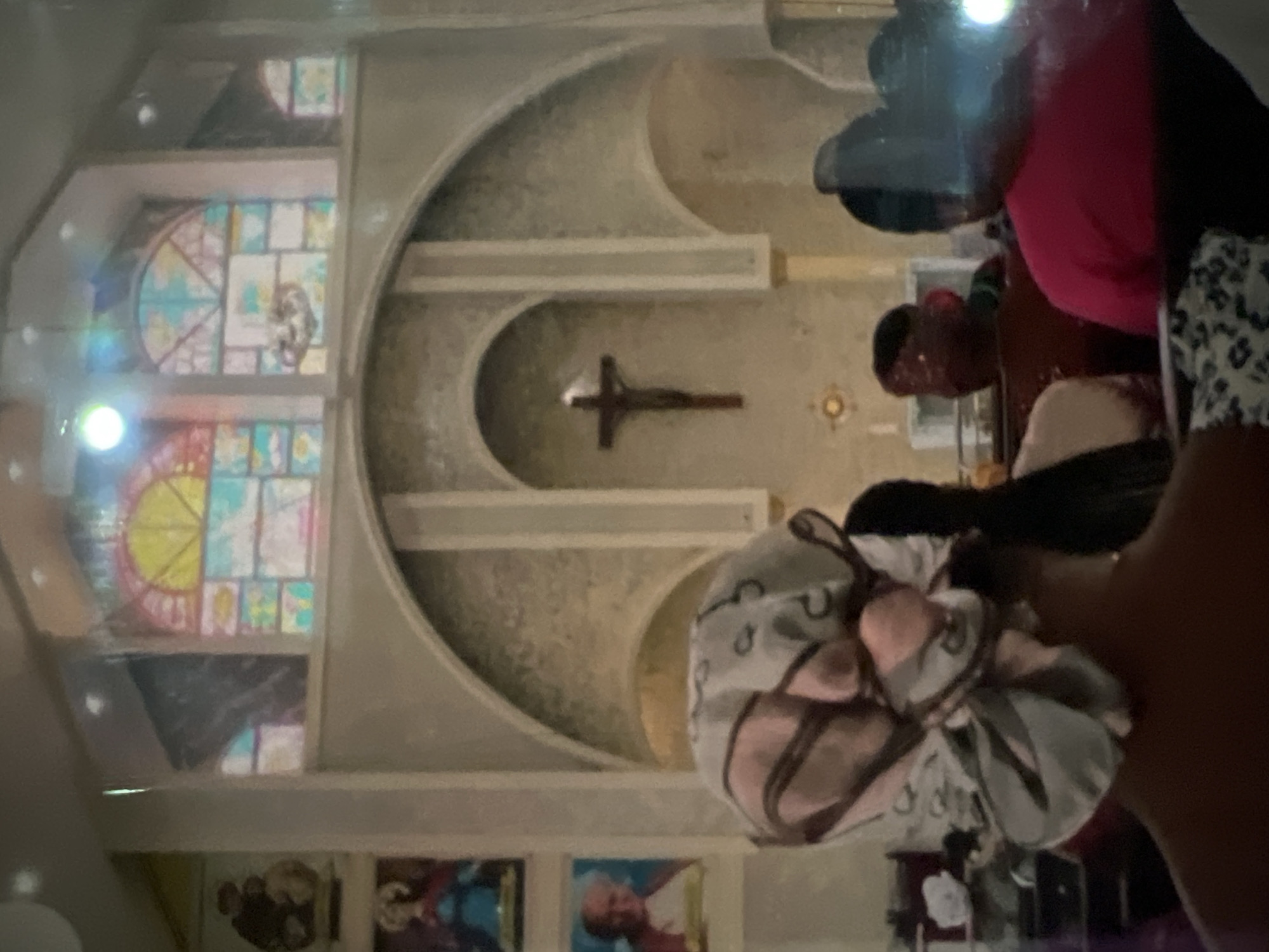 This image depicts the interior of a church, likely a Catholic one, given the presence of a crucifix and religious artwork. In the foreground, the back of a person's head is visible, wearing a patterned headscarf in shades of pink, white, and gray. Behind them, other individuals are seated, their faces mostly obscured. The church features arched architectural elements and stained-glass windows in the background, some of which are illuminated. Religious icons or paintings are displayed on the left wall. The overall lighting suggests it could be daytime or a well-lit interior space. Without further information or visual cues, it is not possible to definitively confirm the location as Lagos, Nigeria, or any other specific city.