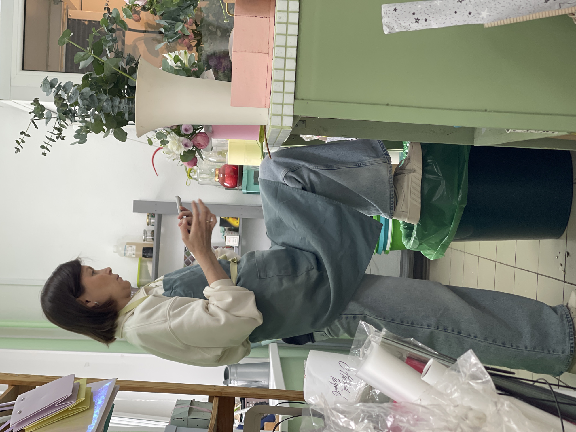 A medium-shot presents a woman with short, dark hair, clad in a white sweater and light blue jeans, seated. She is holding a smartphone in her hands, her attention fixed on the screen. Her facial expression is neutral, suggesting focus or concentration.
She appears to be in a workplace, possibly a florist shop, judging by the presence of flowers and floral arrangement tools visible in the background. A green-colored table is in close proximity, topped with floral materials, and a large trash can is placed near the table's legs. There are shelves in the background holding various items, possibly supplies or decorative objects.
The location appears to be a city due to the constructed environment, but the specific city is Unknown, Unknown, due to the lack of identifiable landmarks or signage.