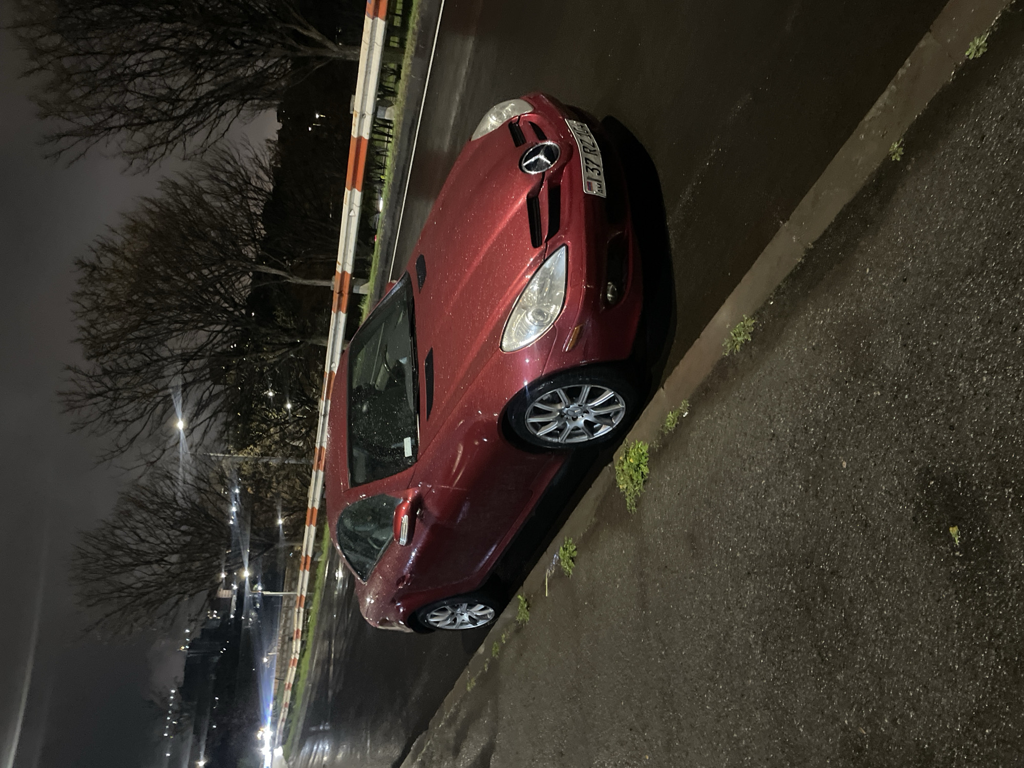 A red Mercedes-Benz SLK is parked on the right side of a wet Yerevan street at night. The car has Armenian license plate numbers beginning with "37 RZ." A concrete curb runs along the street's edge. Bare trees are visible in the background, along with streetlights illuminating the scene. An orange and white striped barrier runs parallel to the street. The street appears to be lightly trafficked.