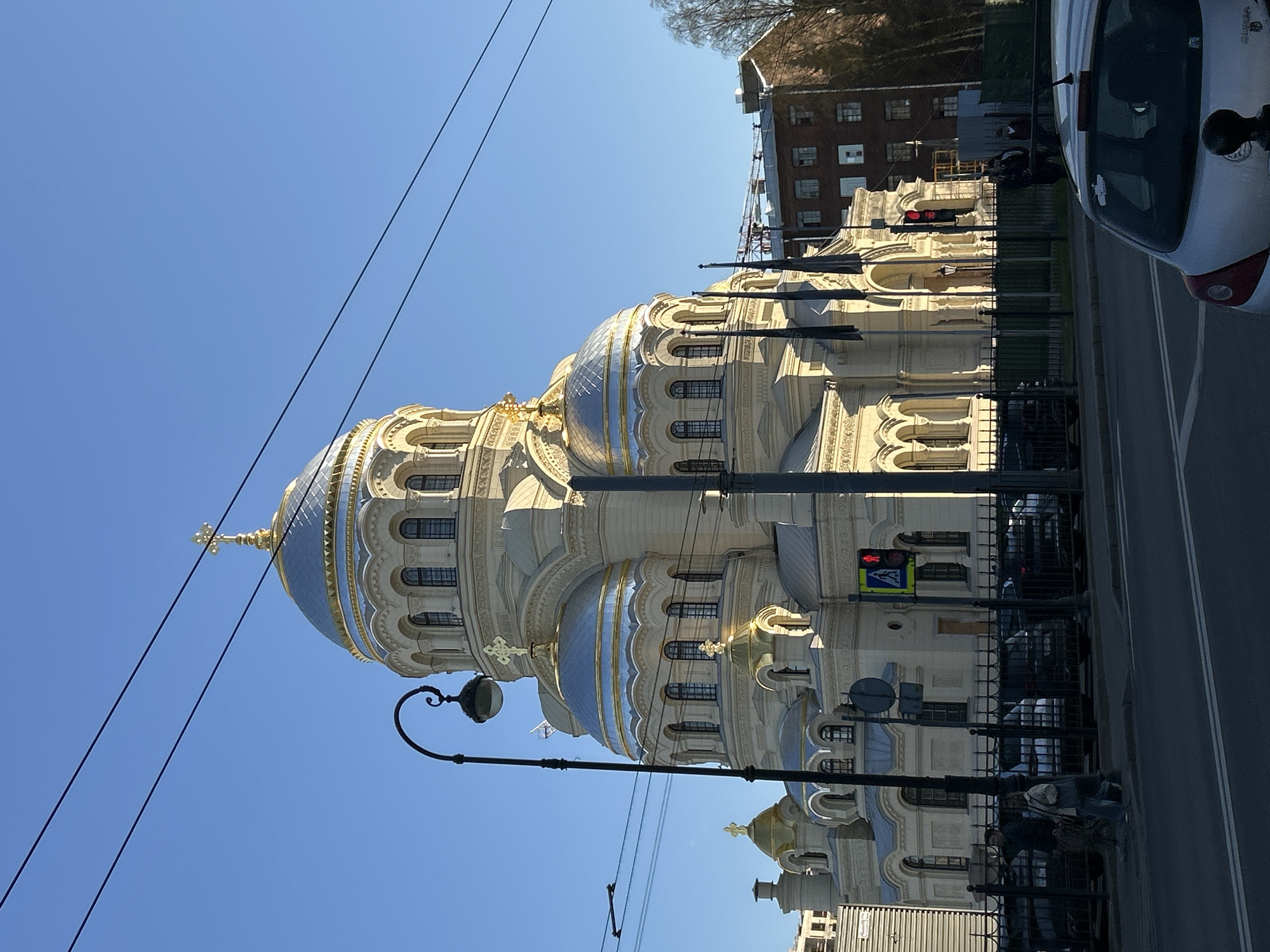 A vibrant, sunny urban scene in Saint Petersburg, Russia, features the magnificent Naval Cathedral of St. Nicholas under a clear blue sky. The grand, light-colored church dominates the central and right portions of the frame, showcasing its distinctive Neo-Byzantine architecture. Multiple domes, primarily a striking light blue adorned with golden accents and ribs, crown the structure. The largest dome is topped with a golden Orthodox cross. The church facade is rich with intricate details, including numerous arched windows and decorative elements, all basking in bright sunlight.
In the foreground on the left, an ornate black street lamp curves upwards, with thin black power lines stretching across the upper left sky. A black metal fence runs horizontally across the lower part of the image, separating the church grounds from the adjacent paved street. On the right side, a white car is partially visible, either parked or driving along the street; legible letters "CYBO" are discernible on its rear, suggesting a car brand or model. Further along the street, a red pedestrian traffic light with a visible red walking figure is present. In the background, behind the cathedral, other urban buildings and distant sparse trees can be seen. The scene is brightly lit, with strong sunlight casting distinct shadows and highlighting the architectural grandeur. No specific activity is depicted, presenting a static yet impressive urban landscape.