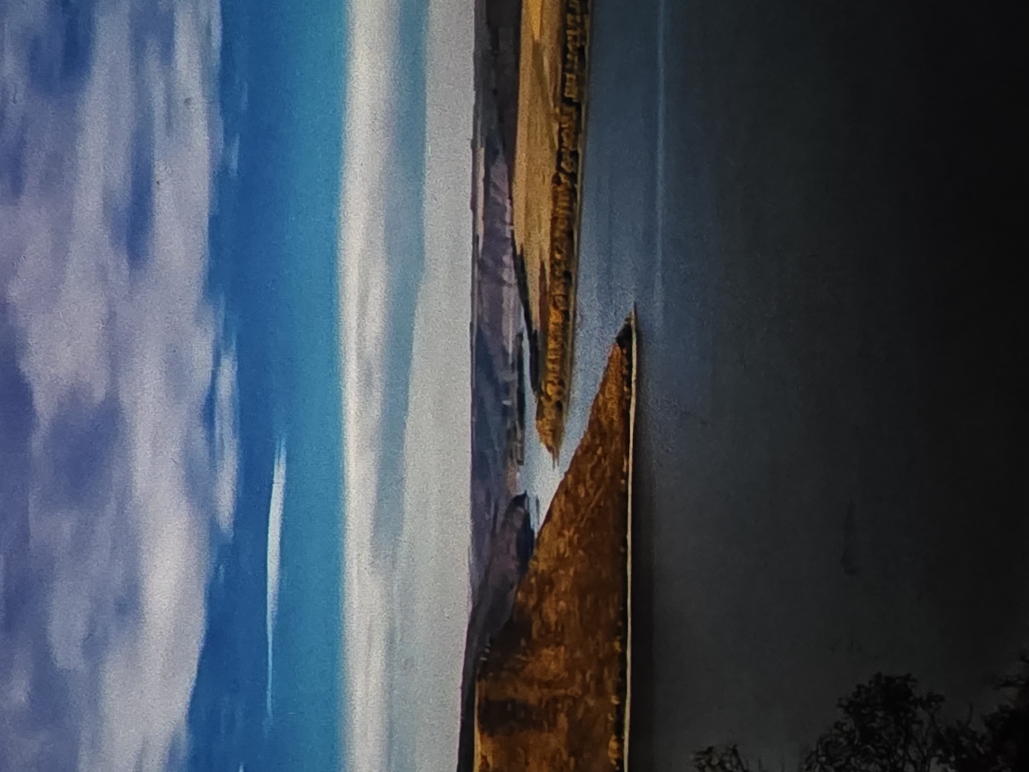 The image depicts a vertical panorama of a landscape. On the left, a bright blue sky with soft, white clouds fills the frame. Moving to the right, there's a transition to a rocky, mountainous terrain in shades of brown and gold, with a deep canyon or valley visible. The texture of the rock suggests a dry, possibly arid environment. Further to the right, the scene darkens into a deep gray or black area, with the silhouette of trees visible at the very bottom, indicating a potential forest or vegetation in the foreground or background.
The lighting suggests it could be daytime, possibly late afternoon or early evening given the shadows and the colors of the rocks. The overall impression is one of natural beauty and ruggedness. There are no people or specific objects clearly identifiable, making it primarily a landscape shot. No text is visible in the image. The scene evokes a sense of vastness and natural grandeur.
Given the provided location context of Dubno, Ukraine, this landscape could represent a natural formation or a scenic view within that region. The contrast between the sky, the rocky landscape, and the dark foreground creates a visually striking composition.