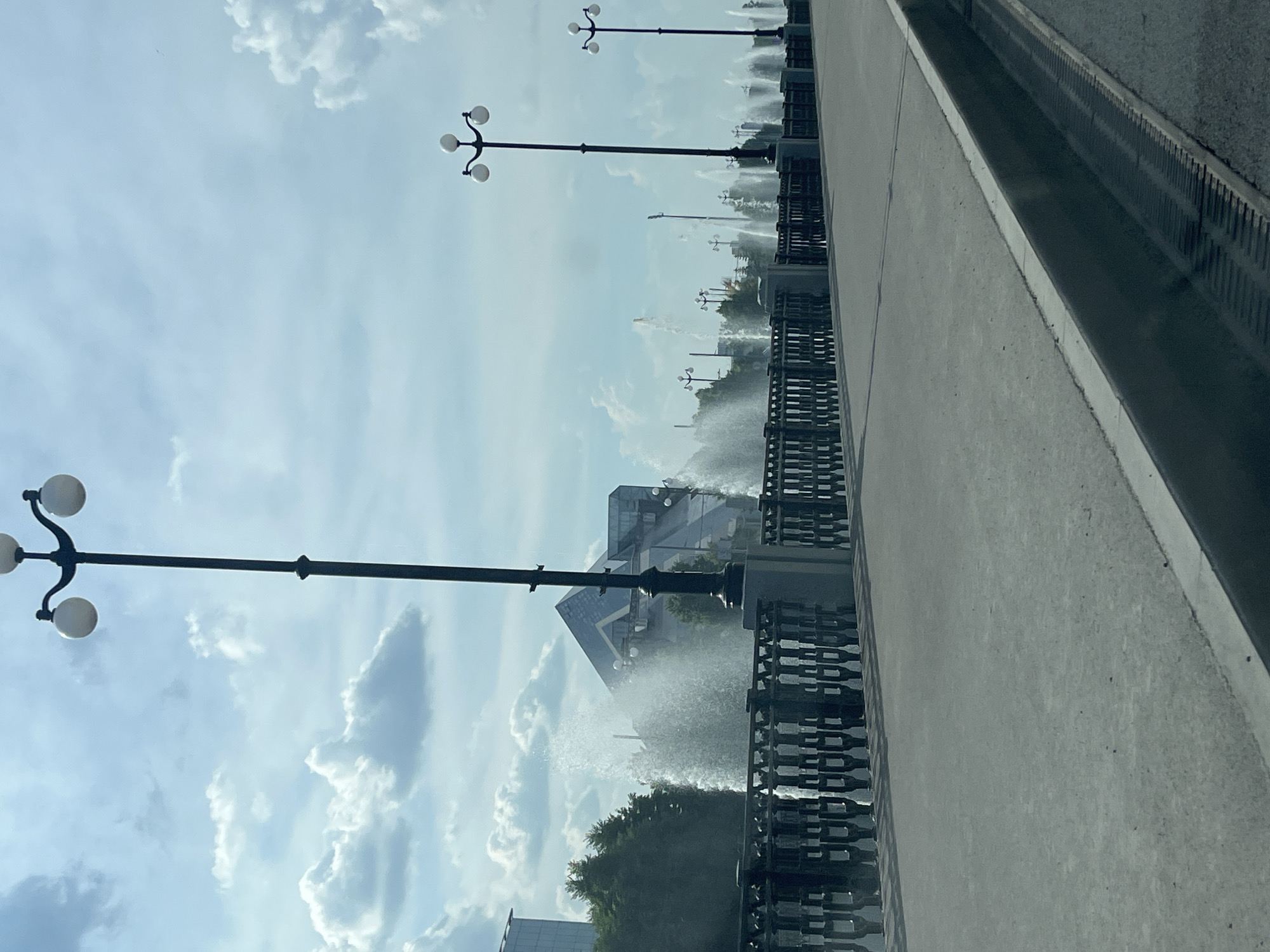 This is a photograph of a fountain on a sunny day, taken from a car. In the foreground, a concrete road and a drainage grate are visible. Beyond that, a paved pedestrian walkway runs alongside a black metal fence. Behind the fence, multiple fountains are spraying water into the air, creating a misty effect. In the background, there are trees, a modern building with a distinctive angular roof, and a clear blue sky with white clouds. The scene is identifiable as the Fountain Ensemble "Kazan" in the city of Kazan, Russia, a popular tourist attraction located on the Kazanka River embankment.