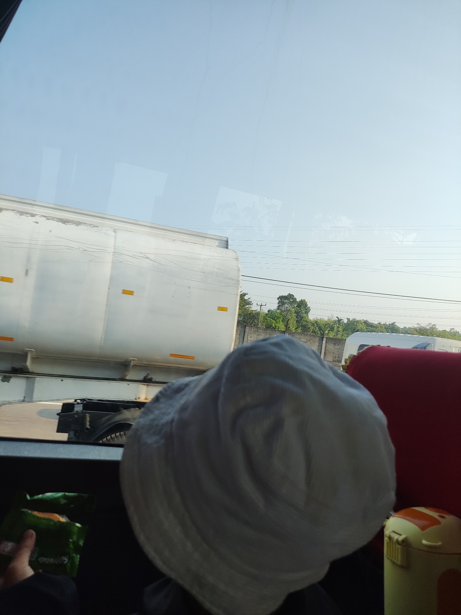 The image is taken from inside a vehicle, looking out a window. The foreground shows the back of a person wearing a white hat. To the right of the hat is a red object, possibly a seat or cushion, and a small yellow container with orange accents. To the left of the hat, a hand is holding a green package of snacks.
Outside the window, a large white tanker truck is visible, with yellow rectangular reflectors on its side. Behind the tanker, there is a concrete wall and lush green trees. The sky is a clear light blue with some faint cloud streaks. Power lines are visible stretching across the sky. The scene appears to be a roadside view, possibly in or around the city of Karawang, Indonesia, given the presence of the tanker truck and vegetation typical of the region.