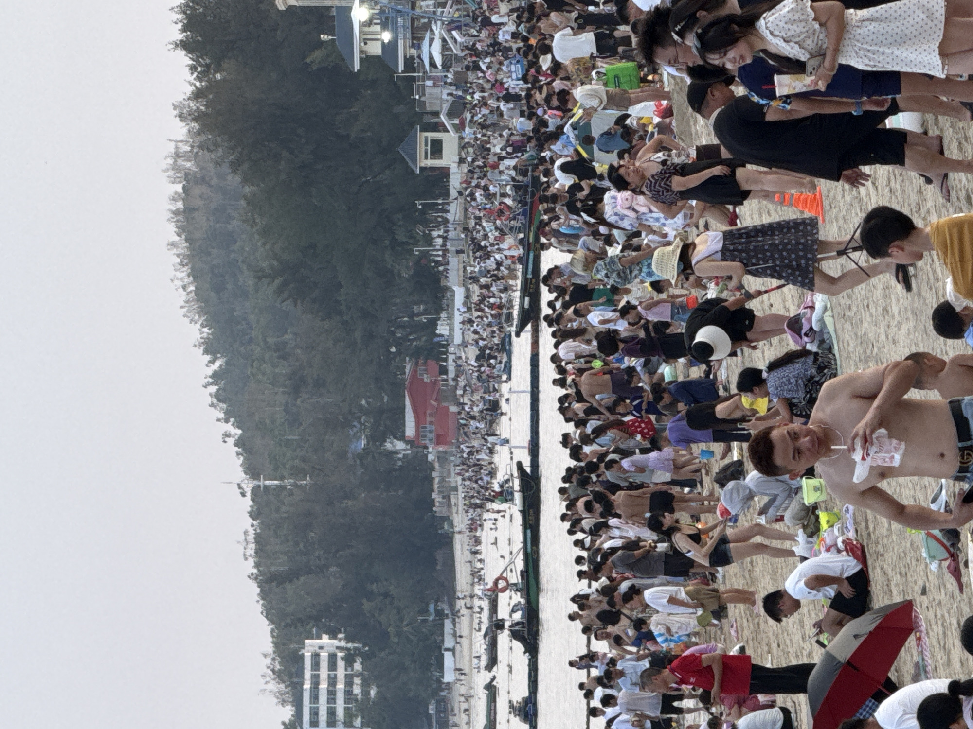 This is a wide-angle, eye-level photograph of a crowded beach in the city of Longgang District, China. The scene depicts a bustling summer day with hundreds of people enjoying the sand and water. The foreground is populated by a dense cluster of beachgoers, many in casual summer attire, with some seated and others standing. A man in the immediate foreground, shirtless, is drinking from a cup with a straw. To his left, a red and black umbrella lies open on the sand.
In the mid-ground, the beach continues to be filled with people, some wading in the shallow water and others gathered on the sand. Several small boats and a larger boat with what appears to be a rescue float are visible in the water. The background reveals a tree-covered hillside with buildings interspersed among the foliage, indicating an urban or semi-urban coastal setting. The sky is overcast and bright, suggesting a warm day. The overall impression is one of a popular recreational area filled with many visitors.