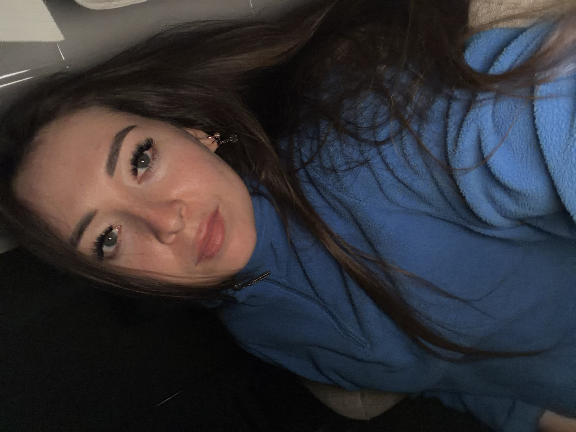 A young woman with long, dark hair and fair skin is shown in a close-up, tilted selfie. She is lying down, wearing a blue fleece jacket with a zipper. Her eyes are open and she is looking towards the camera with a slight smile. Her eyebrows are well-defined, and she appears to be wearing eye makeup and possibly false eyelashes. She has on a small dangling earring in her visible ear. The background is dark and out of focus, but a sliver of white is visible at the top, suggesting an indoor setting. The lighting is dim, possibly indicating nighttime or a dimly lit room. Her lips are slightly parted, revealing a soft pink hue. No text is visible in the image. The overall mood appears relaxed and intimate.