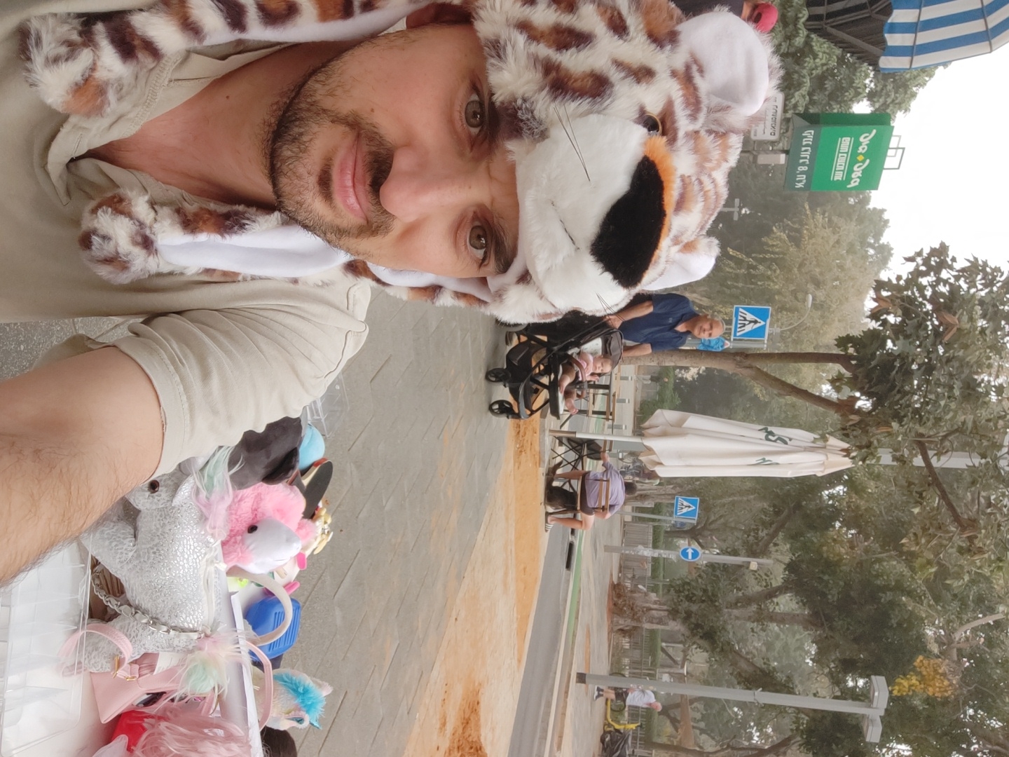 The image is a selfie of a man wearing a faux fur hat resembling a jaguar or similar big cat, with attached ears and a hood. The man is light-skinned with a dark beard and brown eyes. He is wearing a beige or light-colored shirt or hoodie underneath the hat. To his left, in what appears to be a container or bin, there are various soft toys including a unicorn and other colorful plush items.
In the background, there is a cityscape. People can be seen walking or sitting at outdoor cafes. There are traffic signs and trees lining the street. A green and white sign, likely indicating a local business, can be seen in the distance. The architecture and general environment suggest the location is likely in the city of Tel Aviv, Israel.